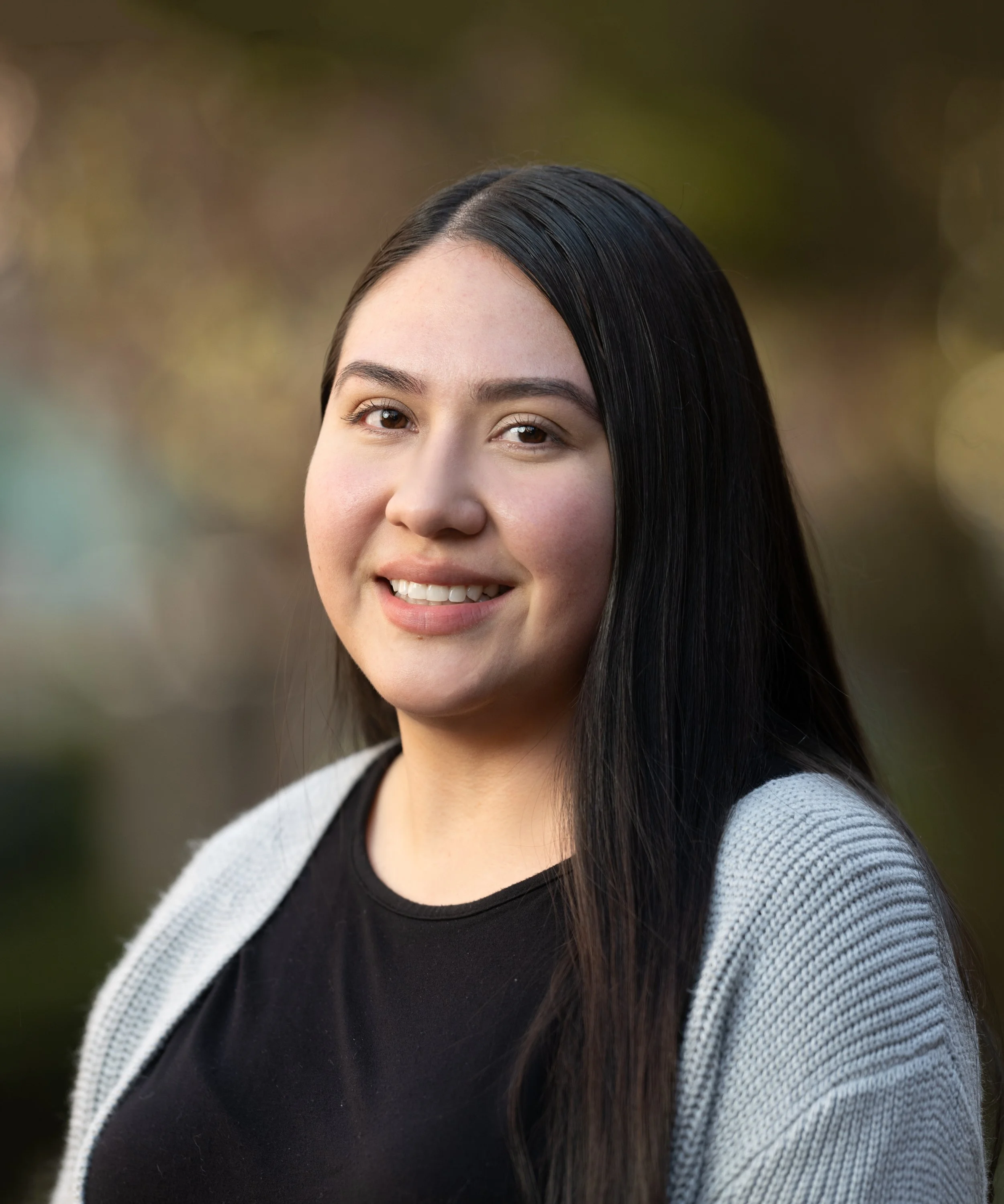 Close-up of a young woman with long, straight black hair, smiling outdoors with a blurred background of trees and foliage.
