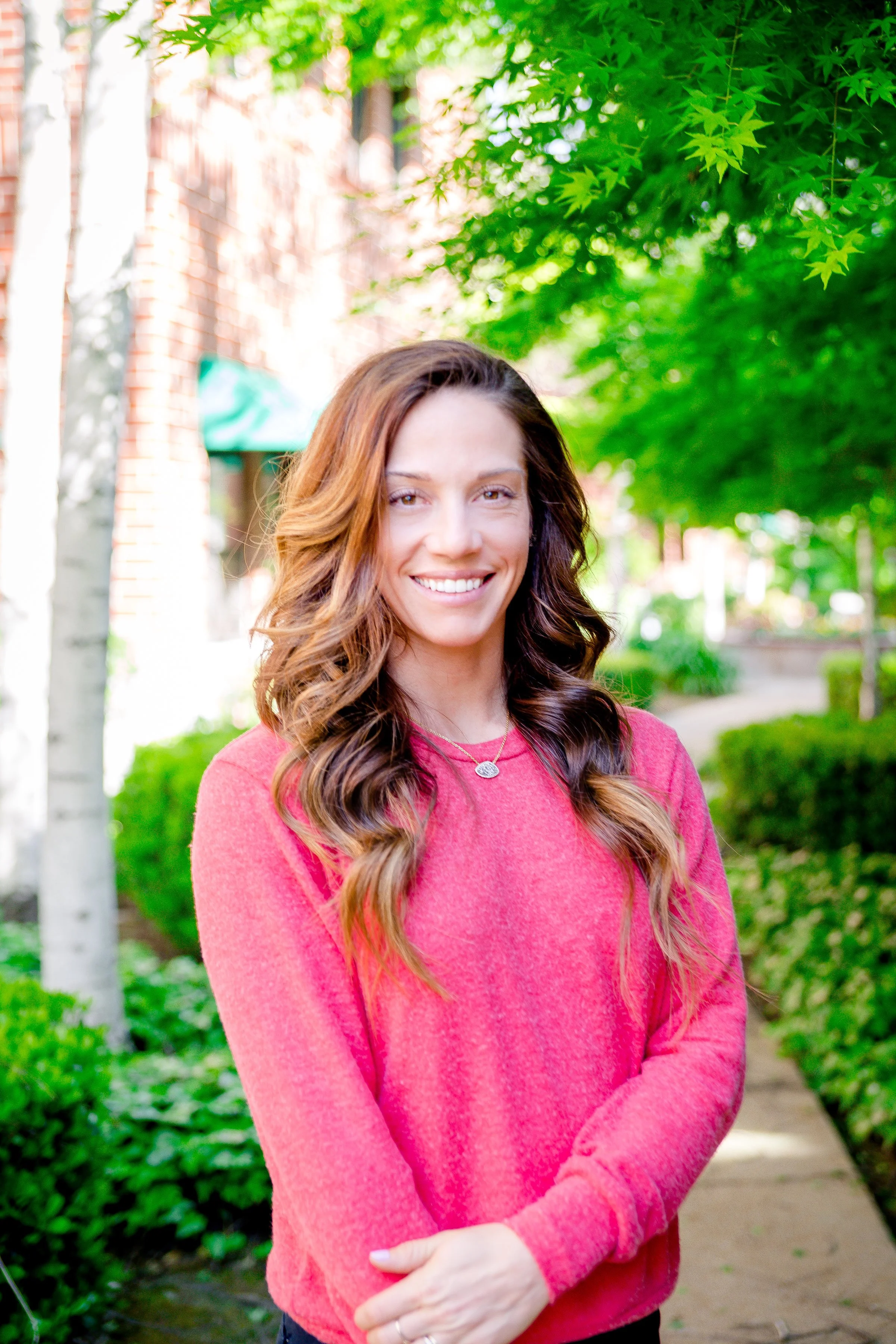 A woman with long wavy brown hair is smiling outdoors, wearing a red sweater, with green trees and a brick building in the background.