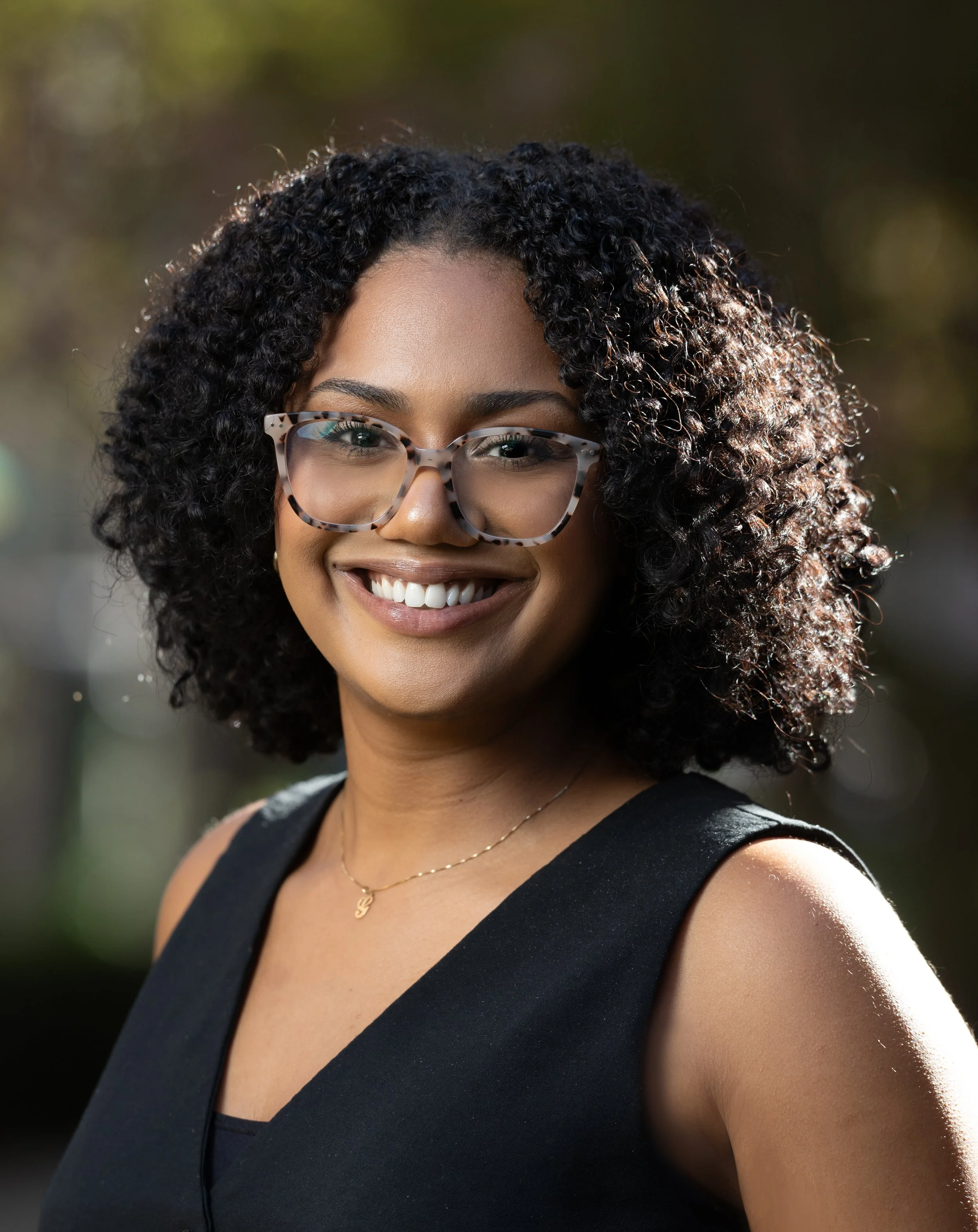 A woman with curly black hair, glasses with patterned frames, smiling, wearing a black sleeveless top and a gold necklace, outdoors with blurred background.