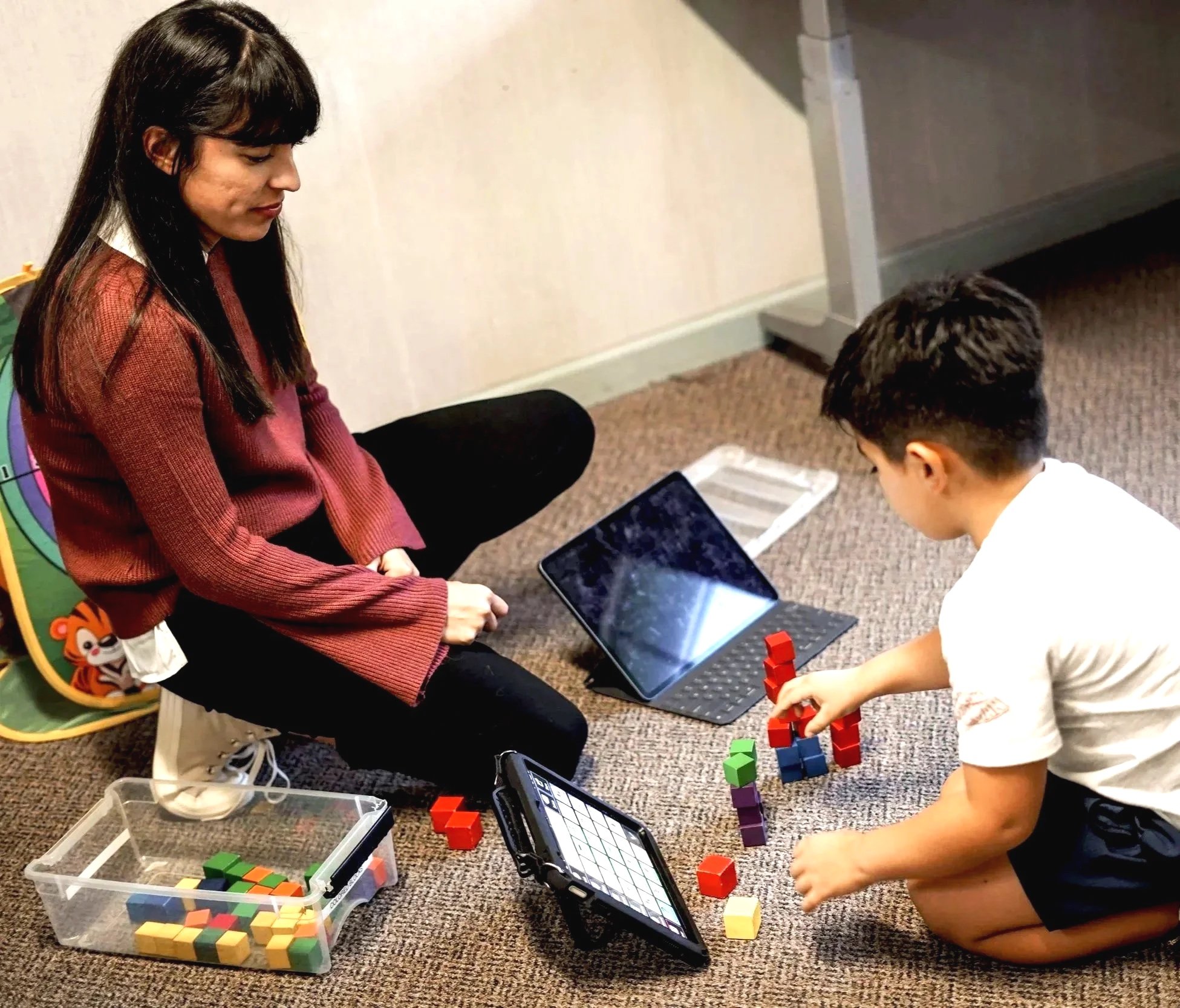 A young woman and a young boy are sitting on the floor playing with colorful blocks and electronic devices, including a tablet and a calculator, in a room with brown carpet and light-colored walls.
