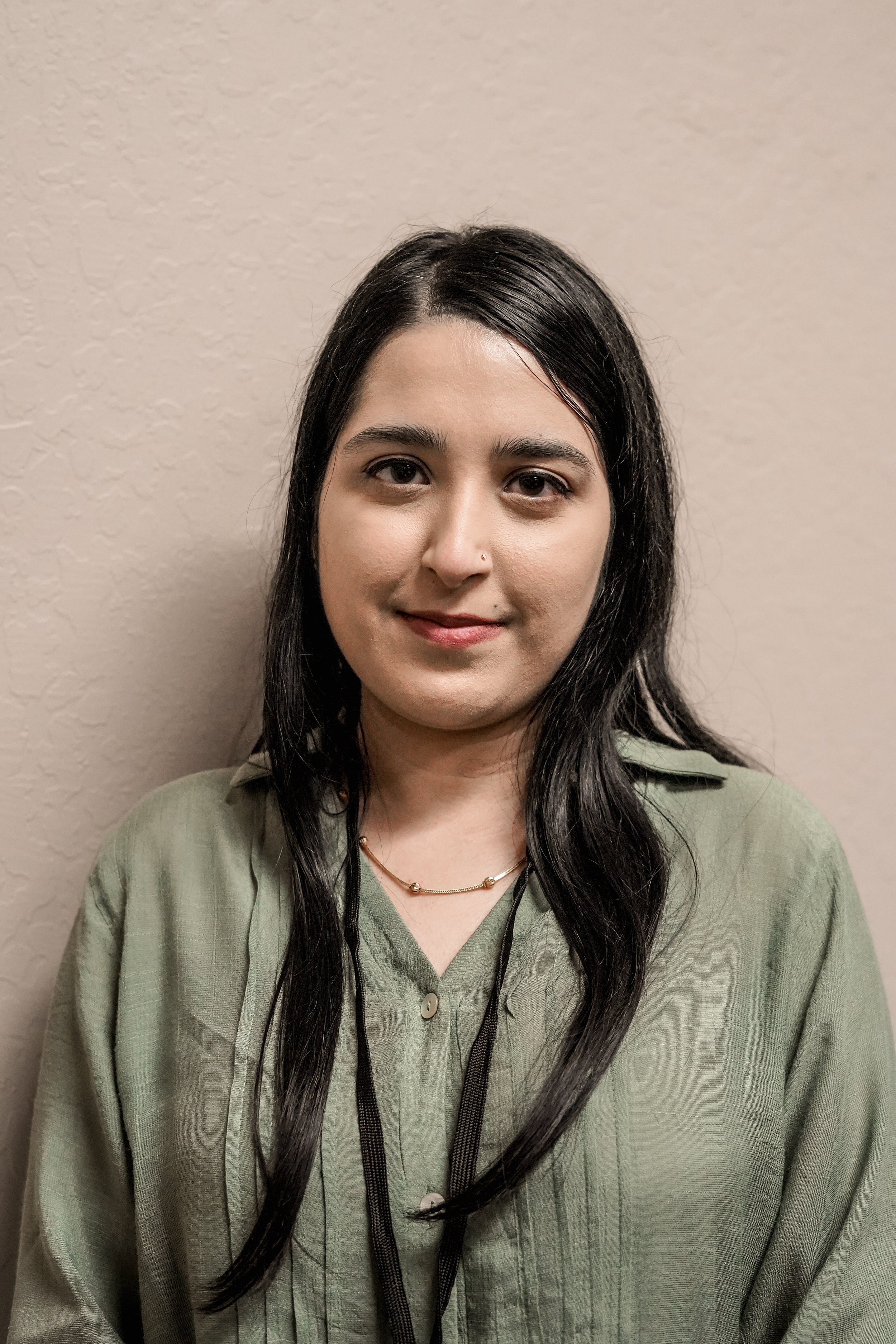 A woman with long dark hair, light brown skin, and wearing a green blouse, standing against a beige wall.