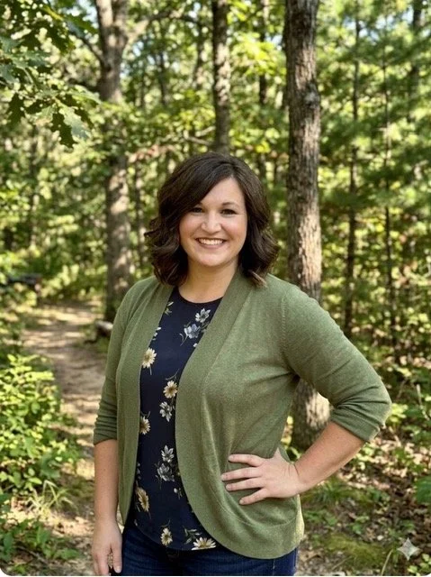 Woman with shoulder-length dark hair smiling, wearing a dark floral top and green cardigan, standing in a wooded area with trees and greenery.