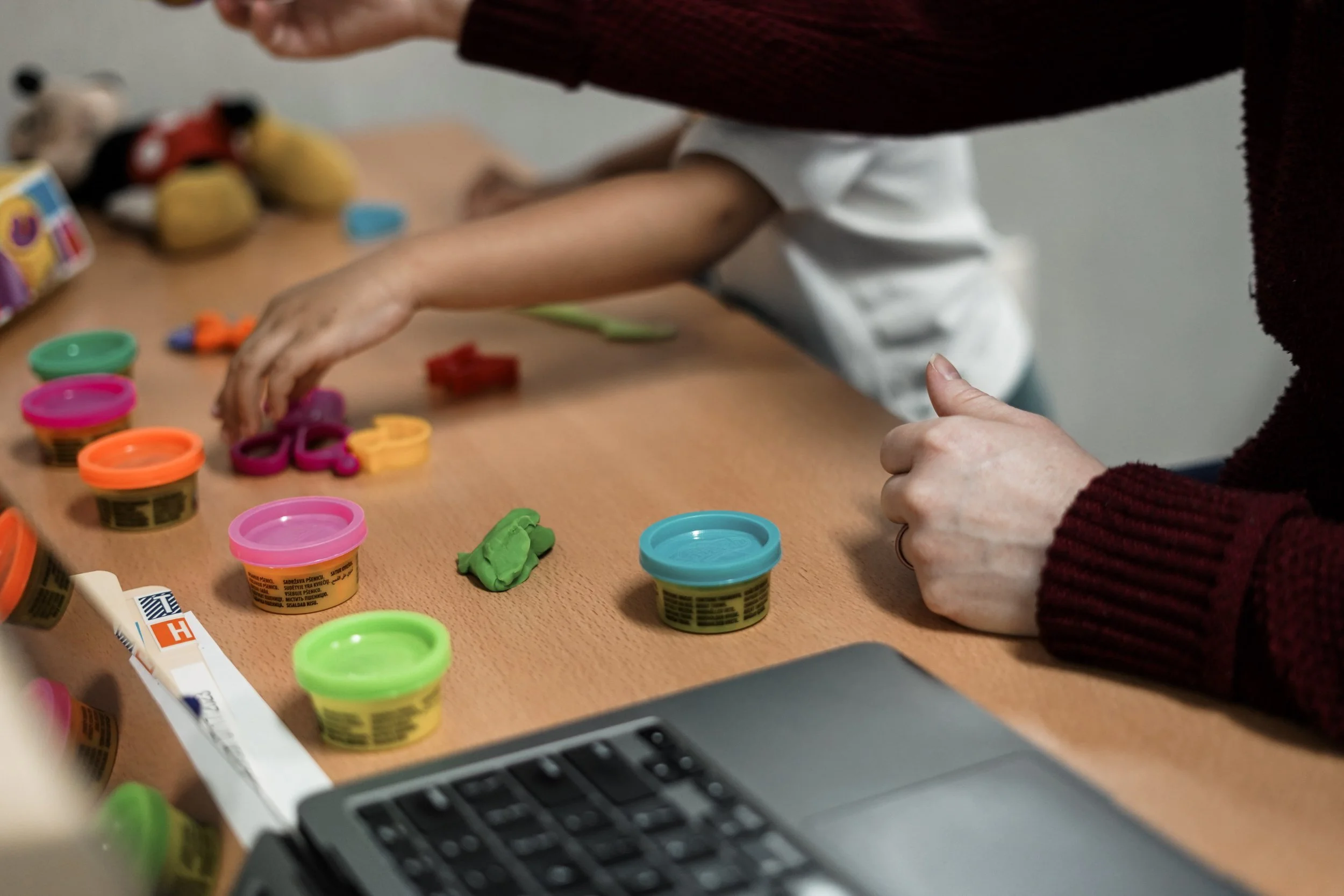 Two people at a table playing with colorful modeling clay, with various containers of clay and some tools scattered on the table, and a laptop partially visible in the foreground.