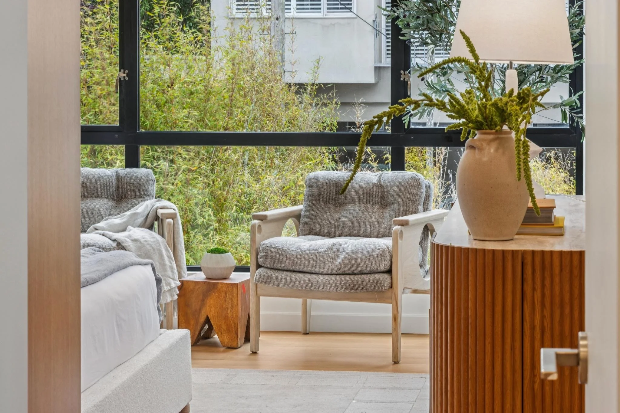 A cozy living room with large windows overlooking greenery. It features light-colored chairs, a white sofa, a side table, and a large ceramic vase with green plants and a lamp on a wooden side table.