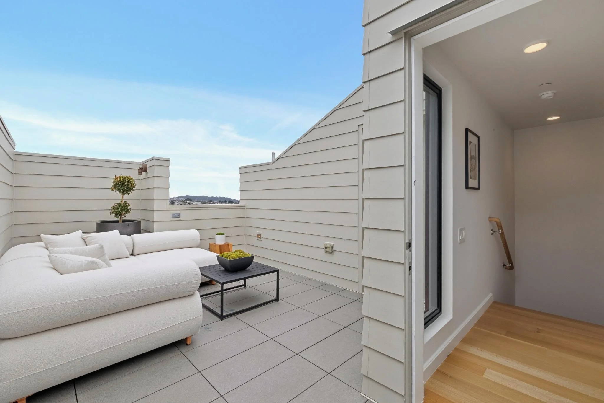 Balcony with white cushioned outdoor sofa, small black table with a black bowl, potted plant, and a view of sky and distant mountains, with part of a house interior visible including a hallway with wooden floor and a framed picture on the wall.