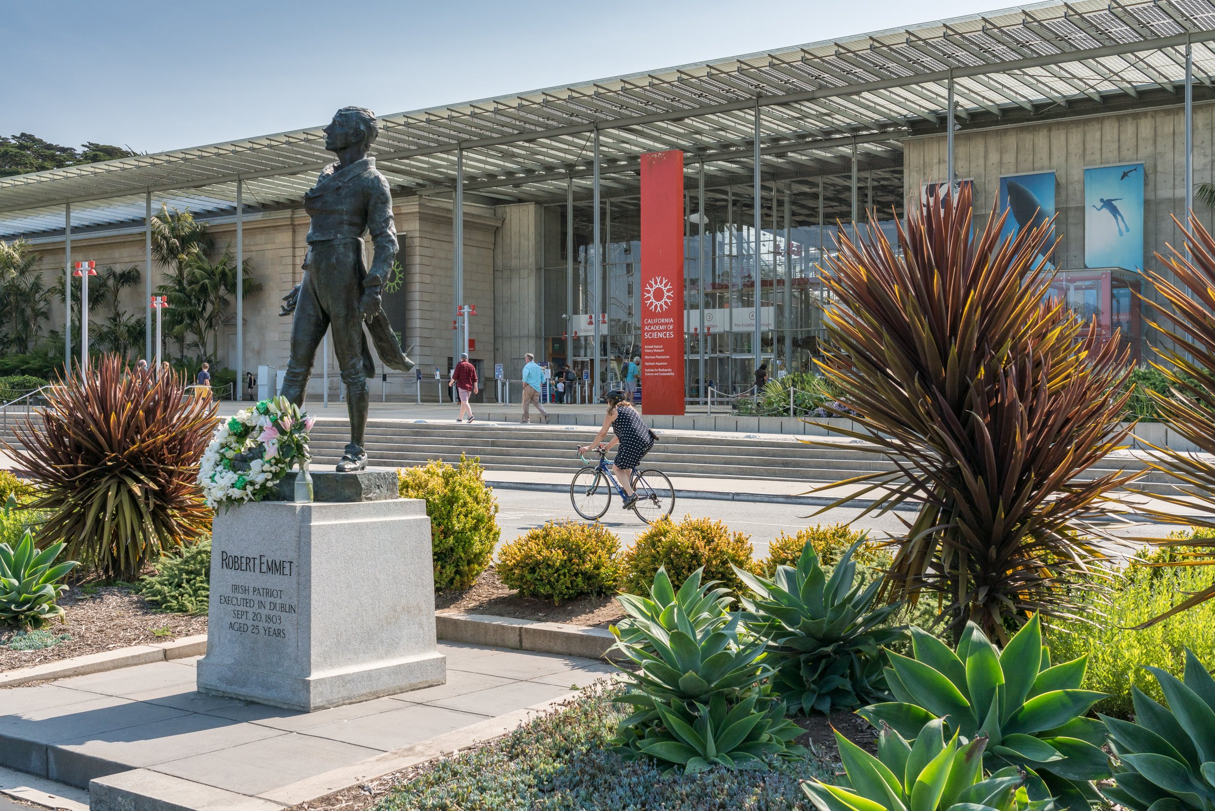 A bronze statue of Robert Emmet, an Irish patriot, stands on a stone pedestal with his name and details inscribed, in a landscaped area with plants. People walk near the entrance of the California Academy of Sciences building in the background, which