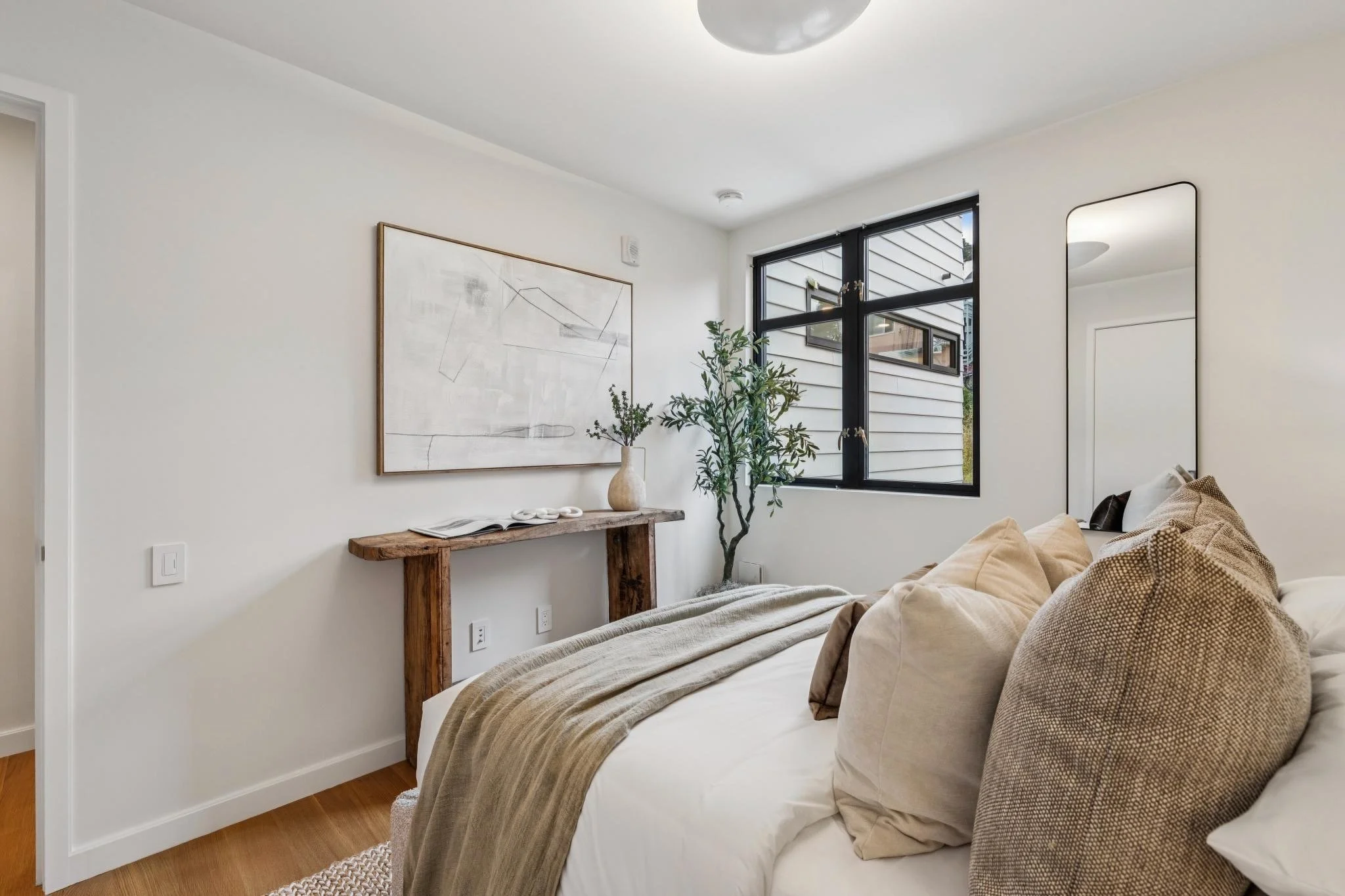 A bedroom with a bed covered in beige and brown pillows and a blanket, a wooden console table with a vase and open book, and decorative plants near a black-framed window. A large abstract artwork hangs on the white wall, and a mirror is visible on th