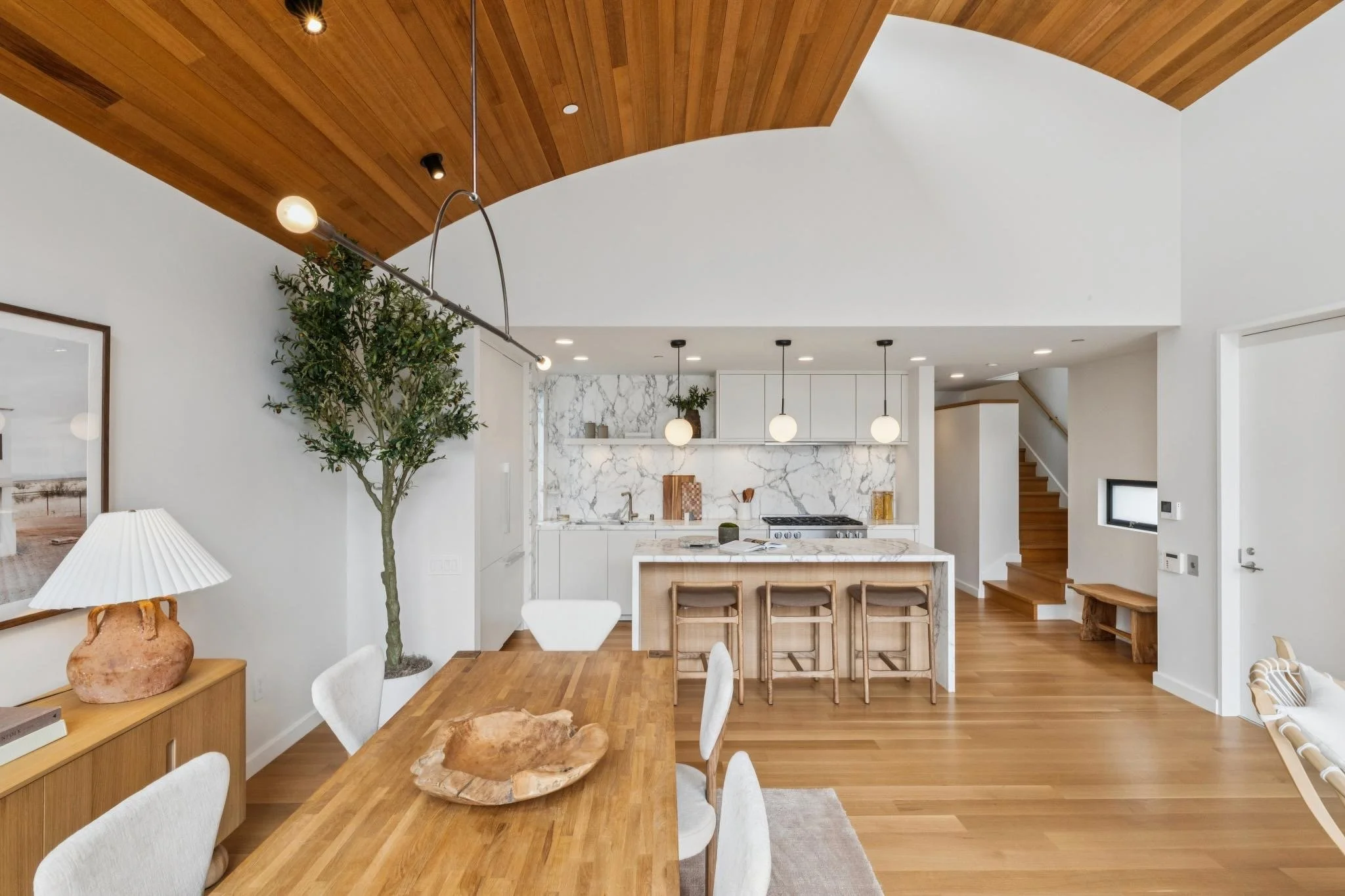 Modern open-concept kitchen and dining area with white walls, wooden floors, and a curved wooden ceiling. The kitchen features white cabinets, marble backsplash, and pendant lighting. The dining table is wooden with white upholstered chairs, and ther