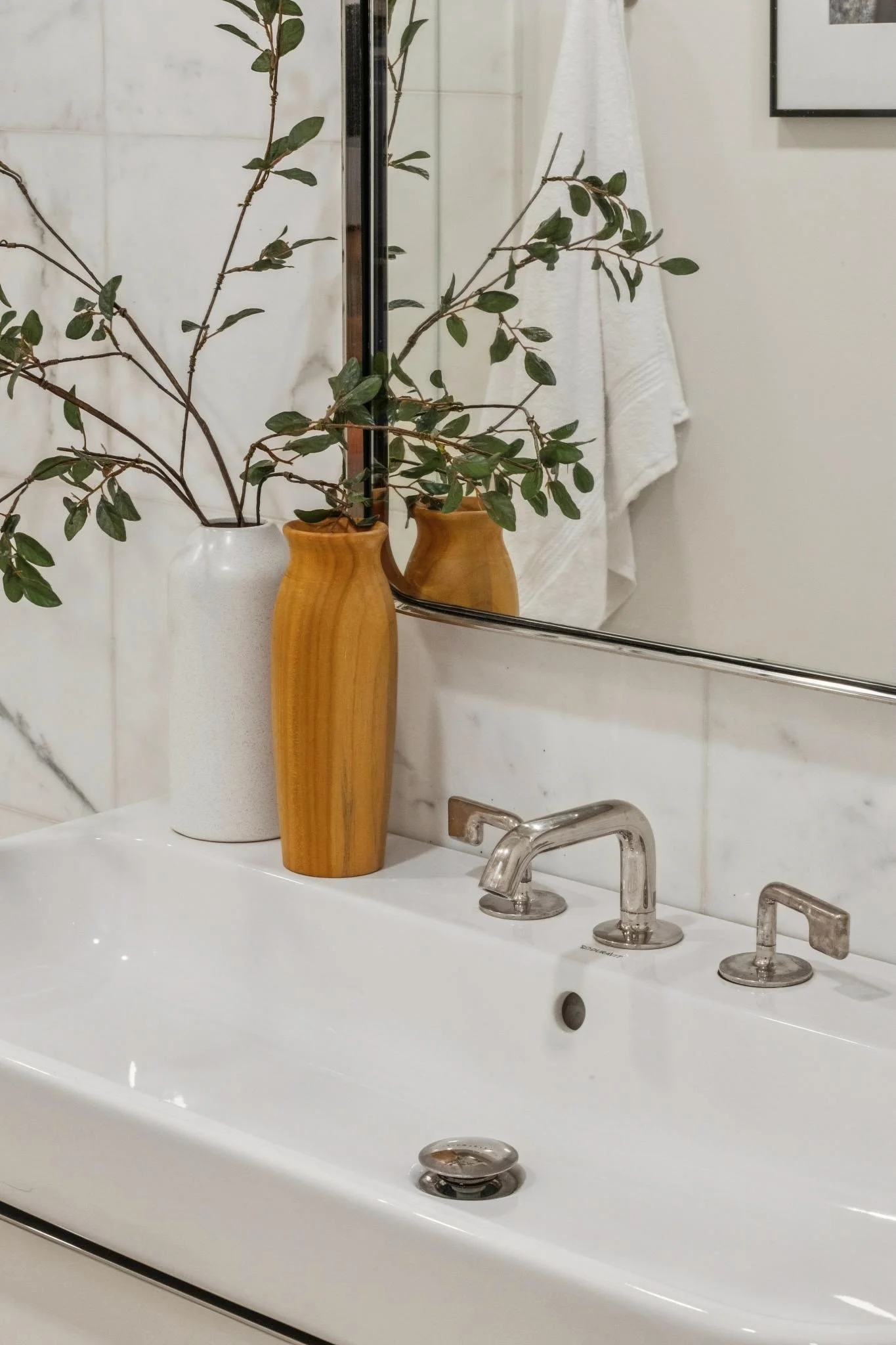 A white bathroom sink with silver faucets and a mirror above, decorated with two vases, one white and one wood-colored, containing green leafy branches, and a white towel hanging on the wall.