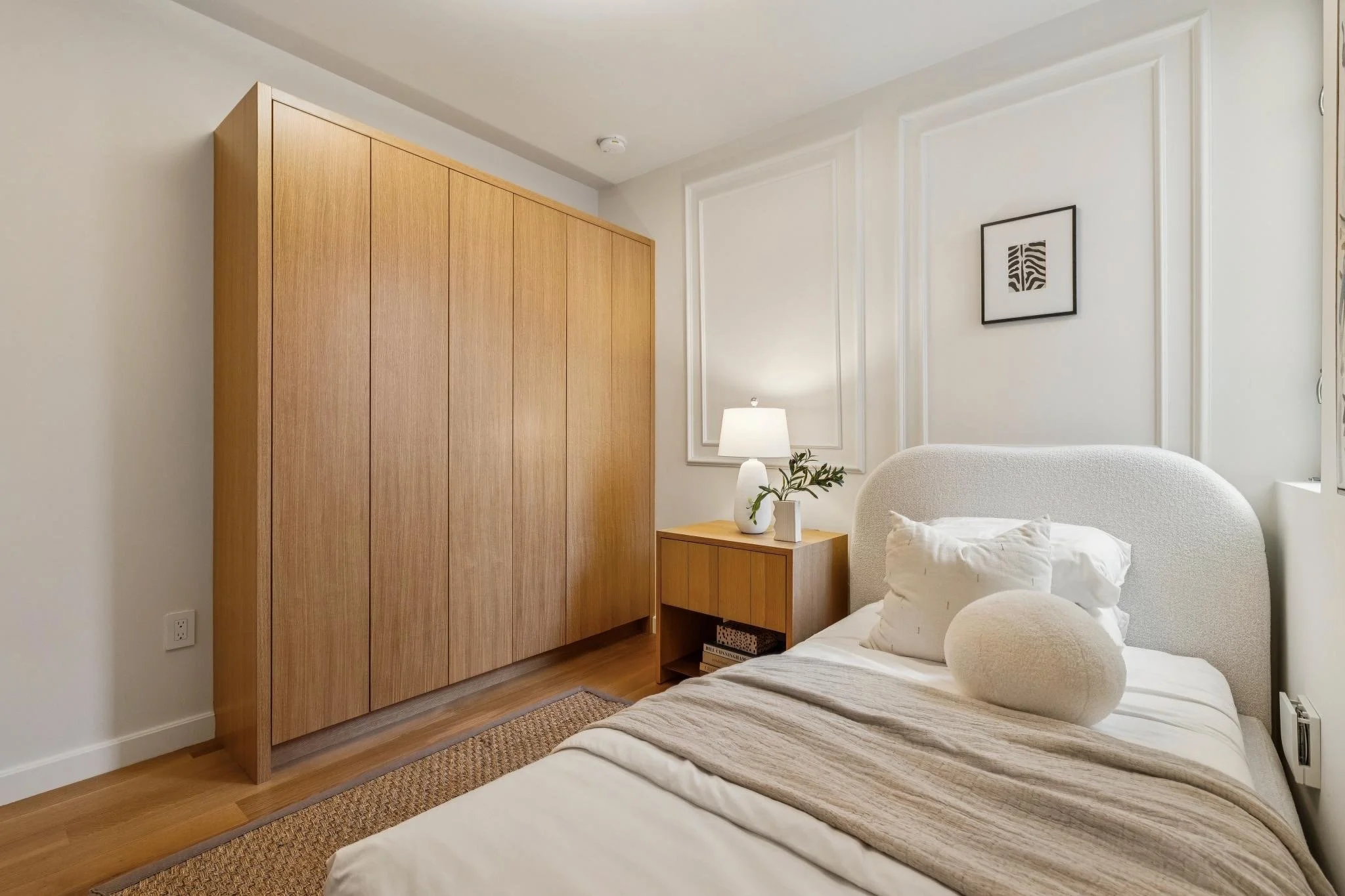 A cozy bedroom corner with a wooden wardrobe, a small bedside table, a white table lamp, a framed abstract art piece on the wall, a white upholstered armchair, and a bed with neutral bedding, all illuminated by natural light.