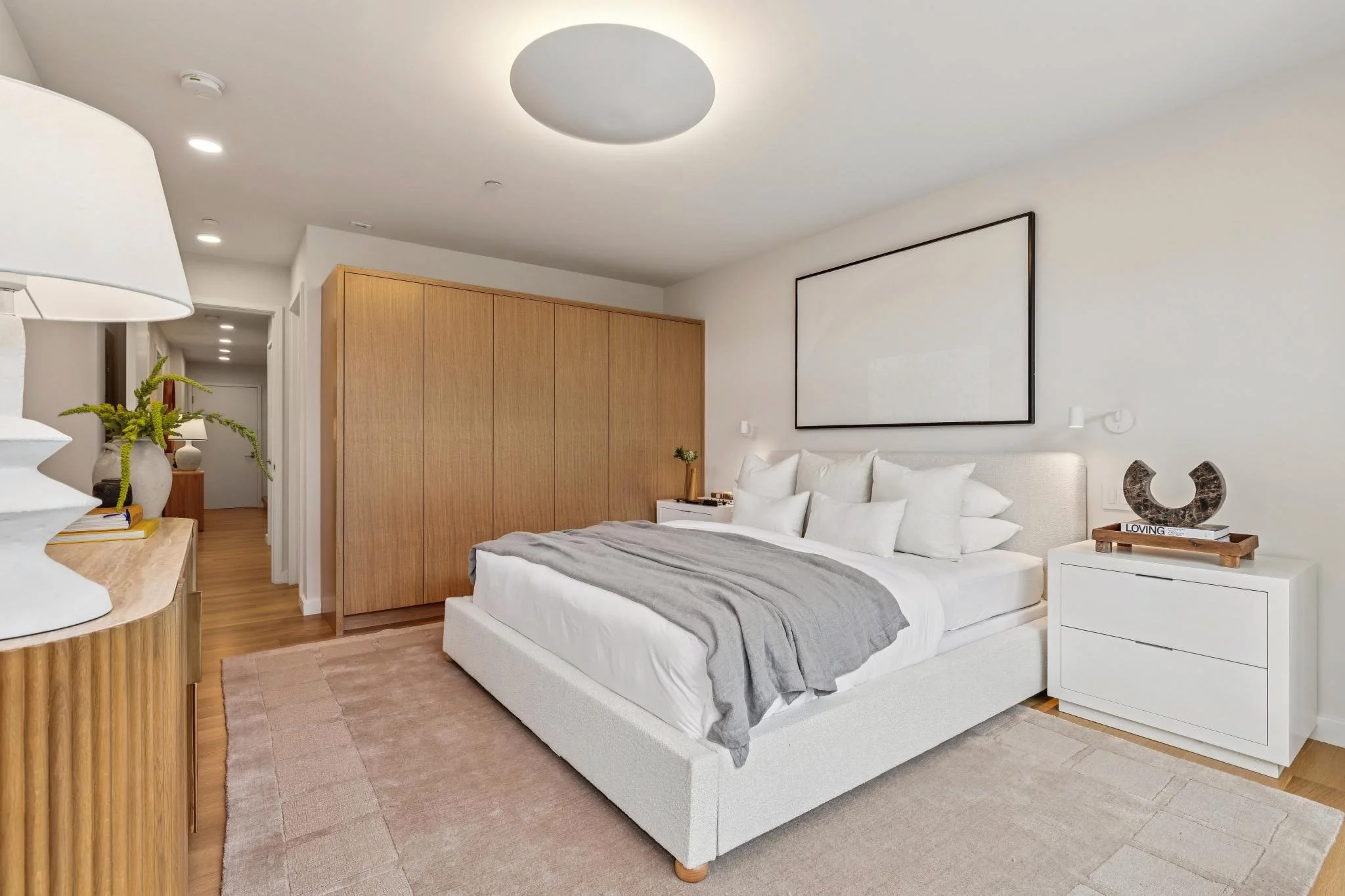 Modern bedroom with white bed, gray blanket, white nightstand, and wooden closet doors. Decorative items include a black sculpture, small plants, and books. Recessed ceiling lights and a large blank black frame on the wall.