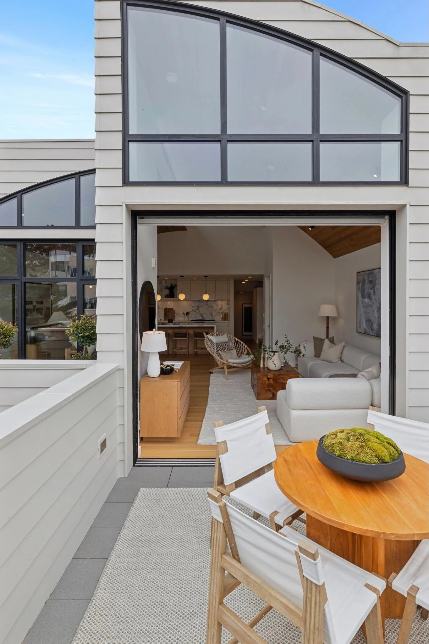 Balcony with wooden table and white chairs, potted plants, view into modern living room with white sofa, wooden coffee table, and kitchen in the background with hanging lights and marble countertop.