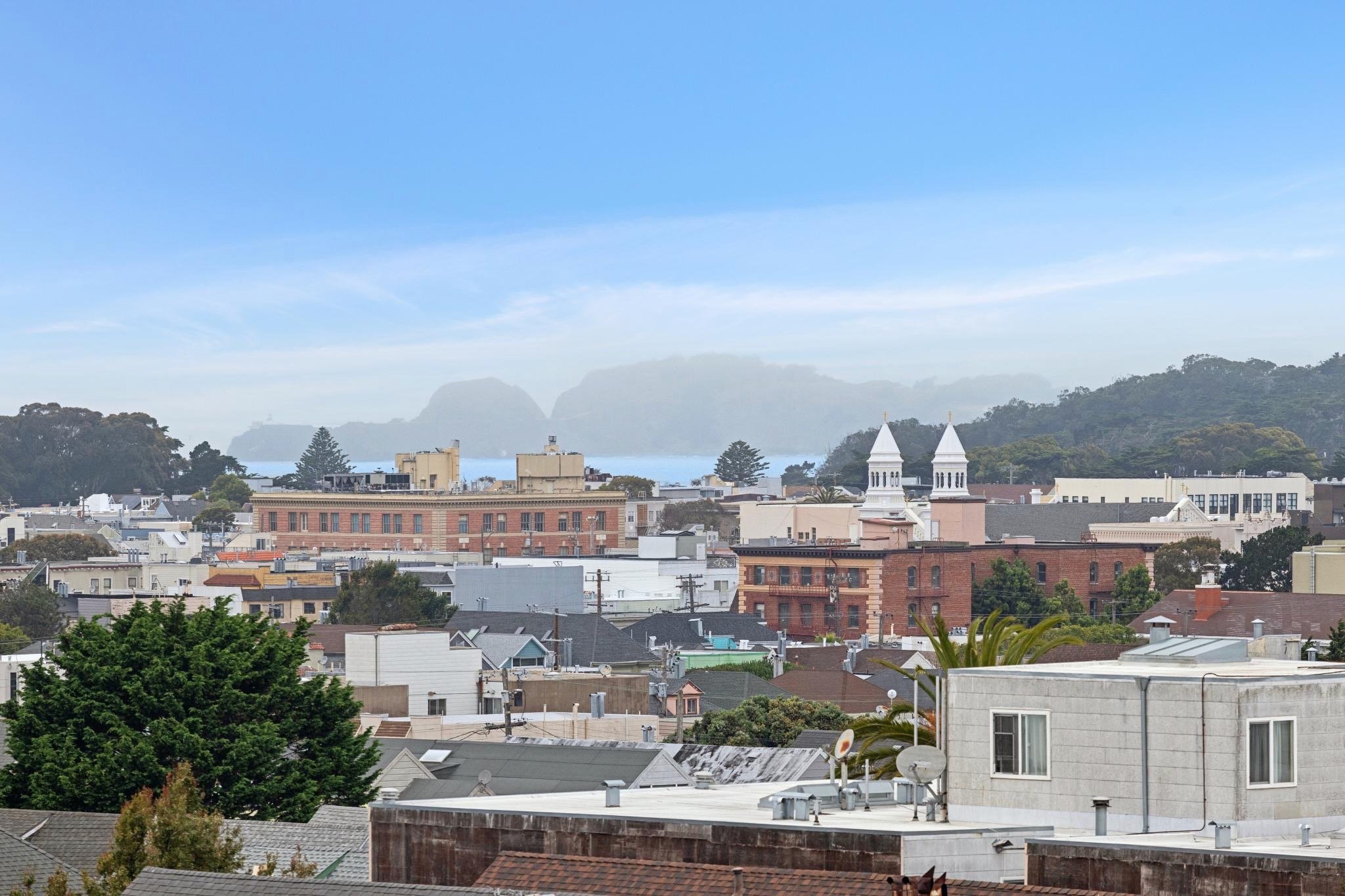 City skyline with buildings, trees, and church steeples, hills in the distance under a blue sky with clouds.