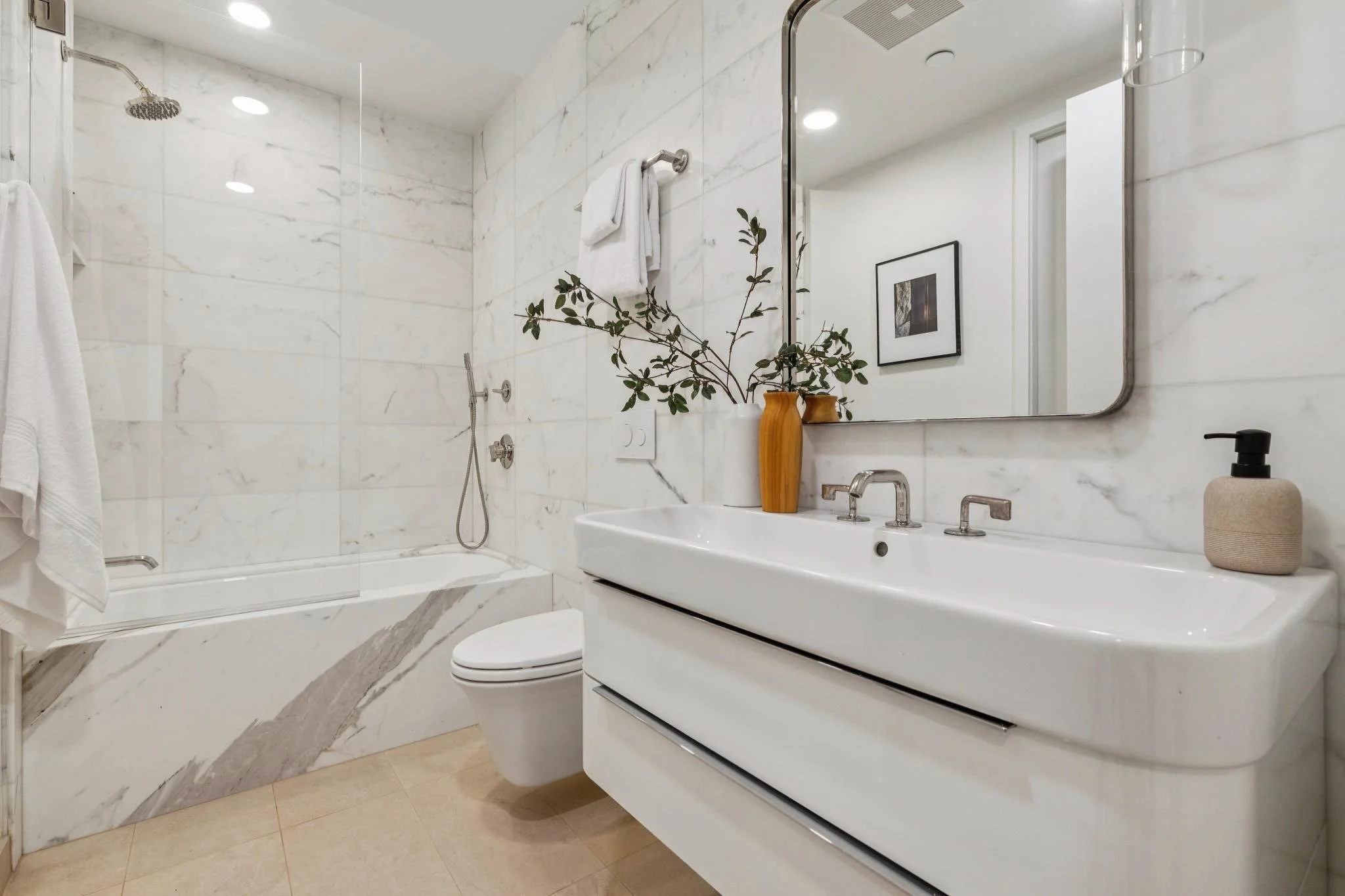 Modern bathroom with white marble walls, a bathtub with a glass shower enclosure, a white vanity with a sink, a large mirror, a potted plant, towels, and bathroom accessories.
