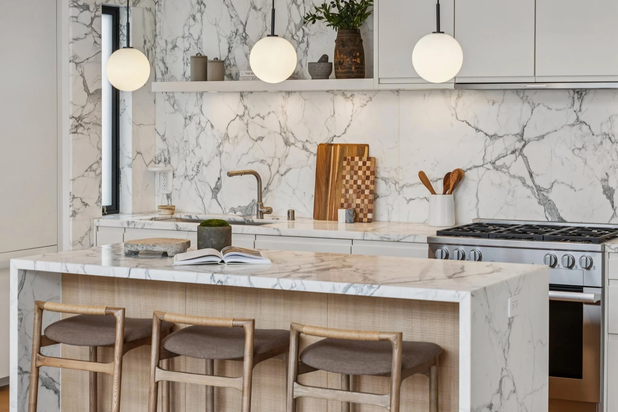 Modern kitchen with marble countertops and backsplash, white cabinets, wooden bar stools, and hanging globe pendant lights.