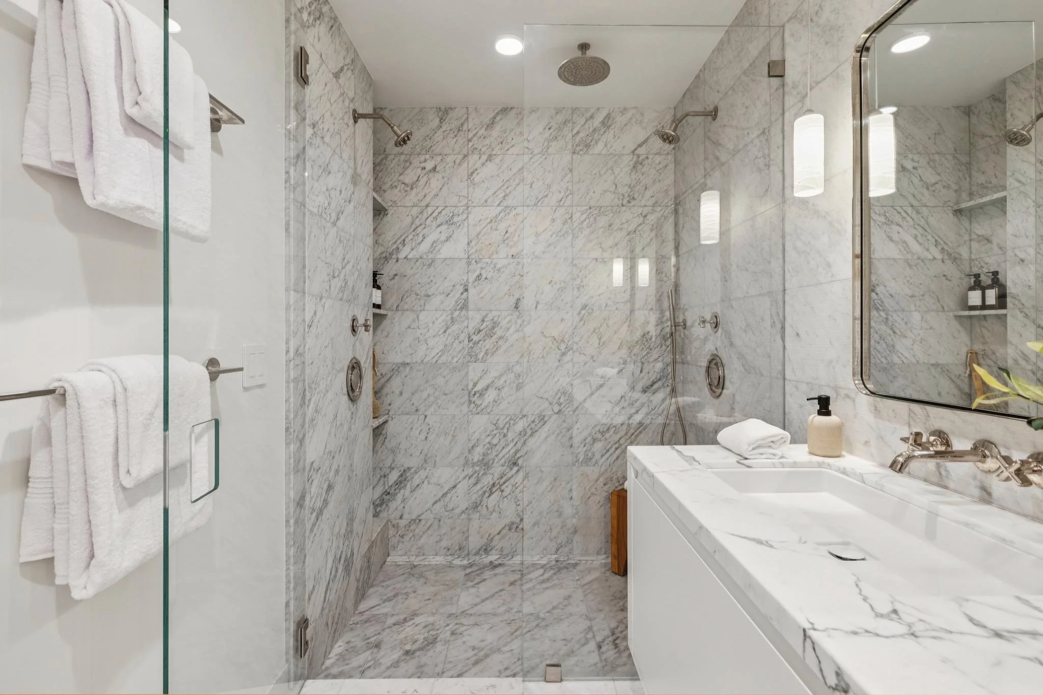Inside a modern shower with marble walls, dual showerheads, and built-in shelves holding toiletries, adjacent to a white vanity with a marble countertop and sink.