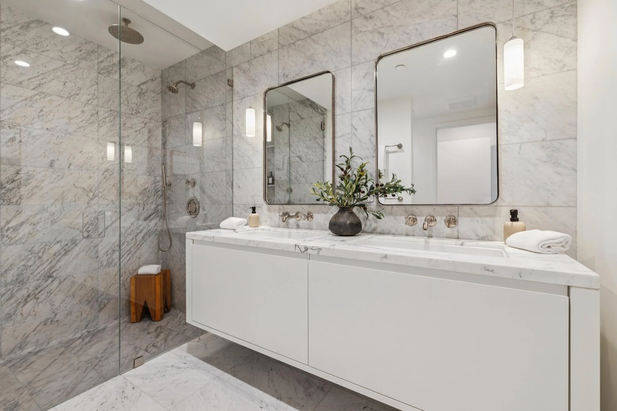 A modern bathroom with a marble shower and vanity. The marble features a white background with gray veining. The shower has a glass door and a rainfall showerhead. The vanity below the large mirror has a white countertop with a potted plant, soap dis