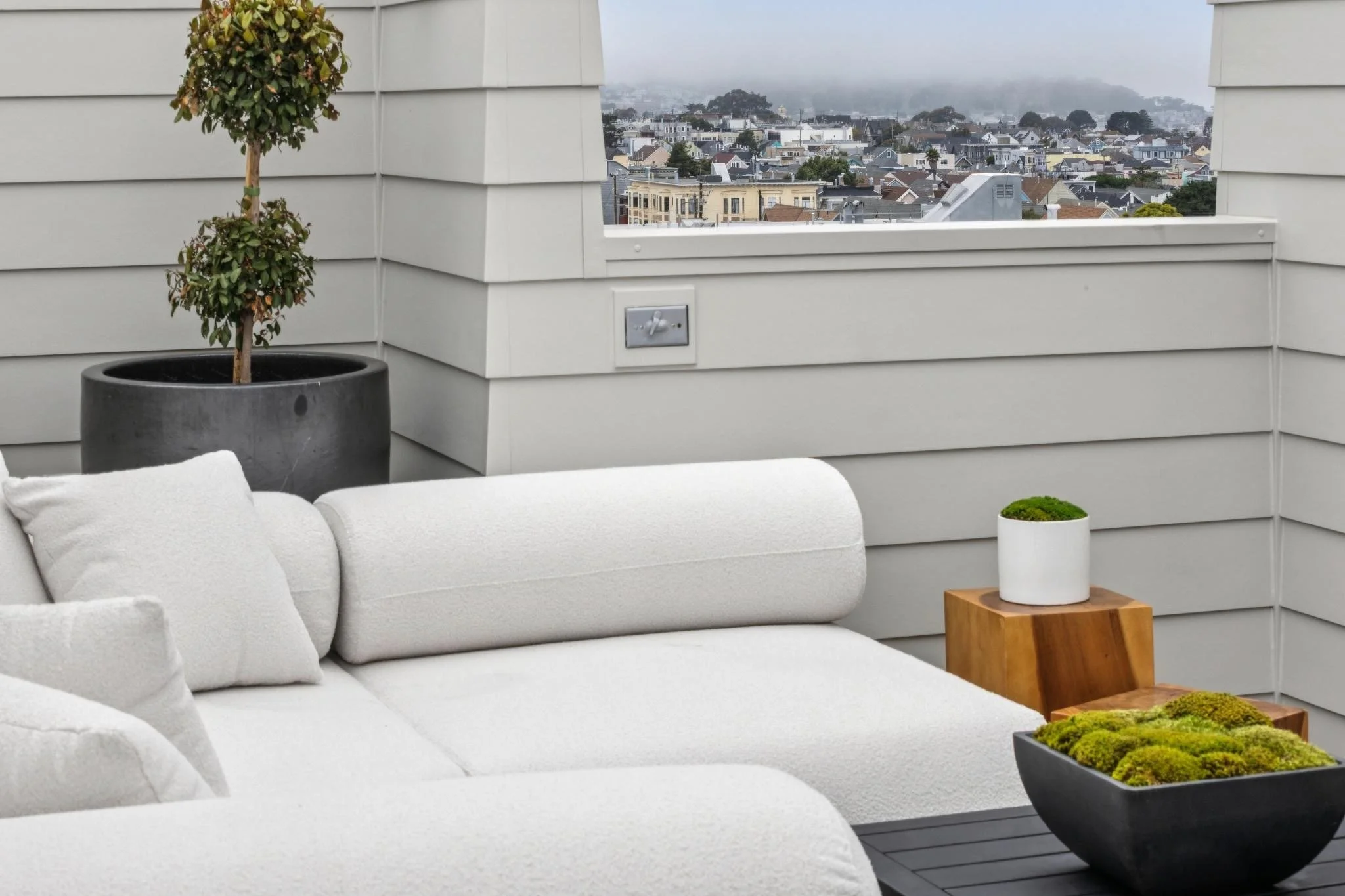 Balcony with white outdoor furniture, potted plants, and a view of a cloudy, foggy cityscape in the background.