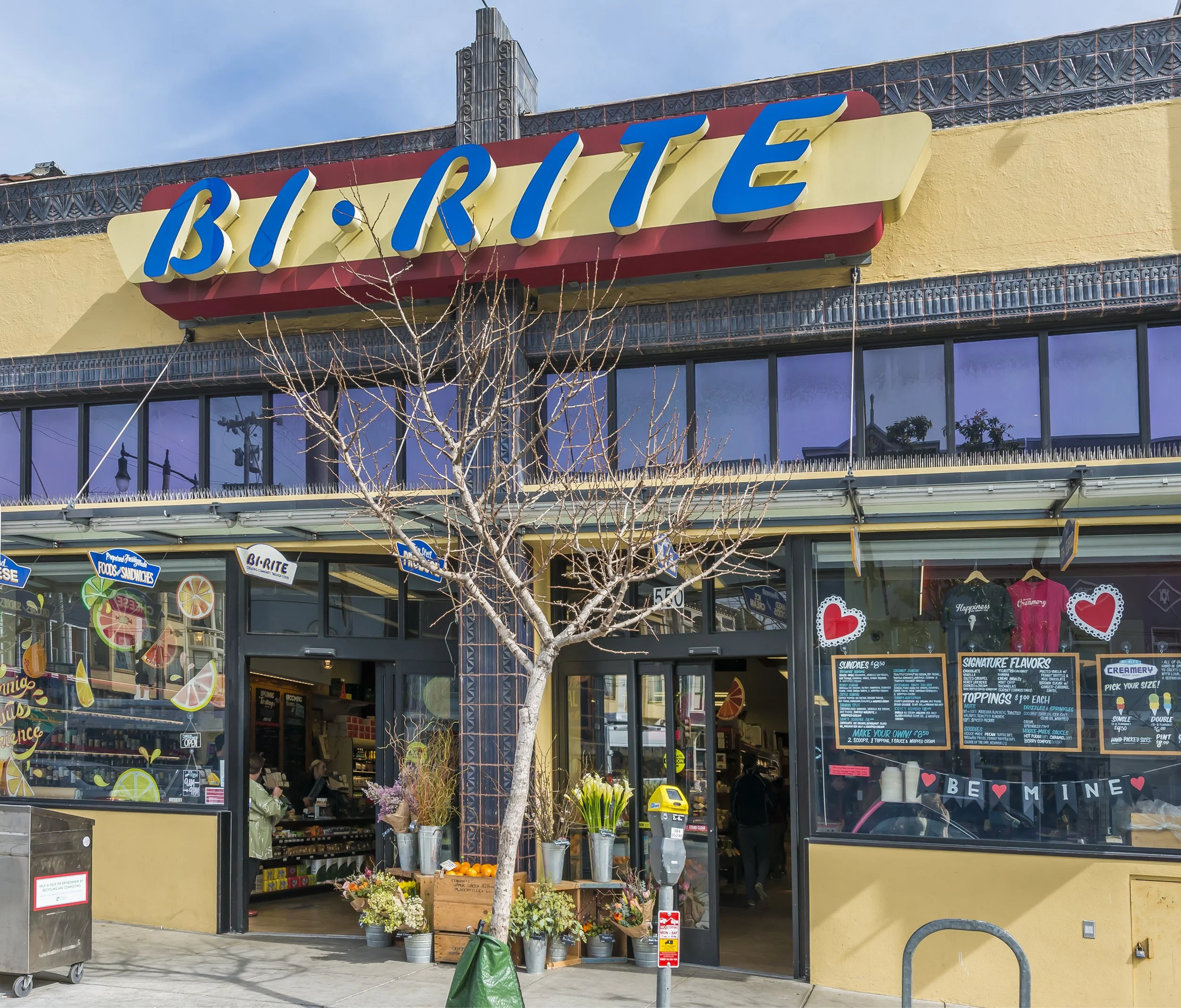 View of a store with a large sign reading 'Brite'. The storefront has glass windows displaying colorful signs and a menu board. Outside, there are potted flowers and a small tree with bare branches. The interior of the store appears to sell ice cream