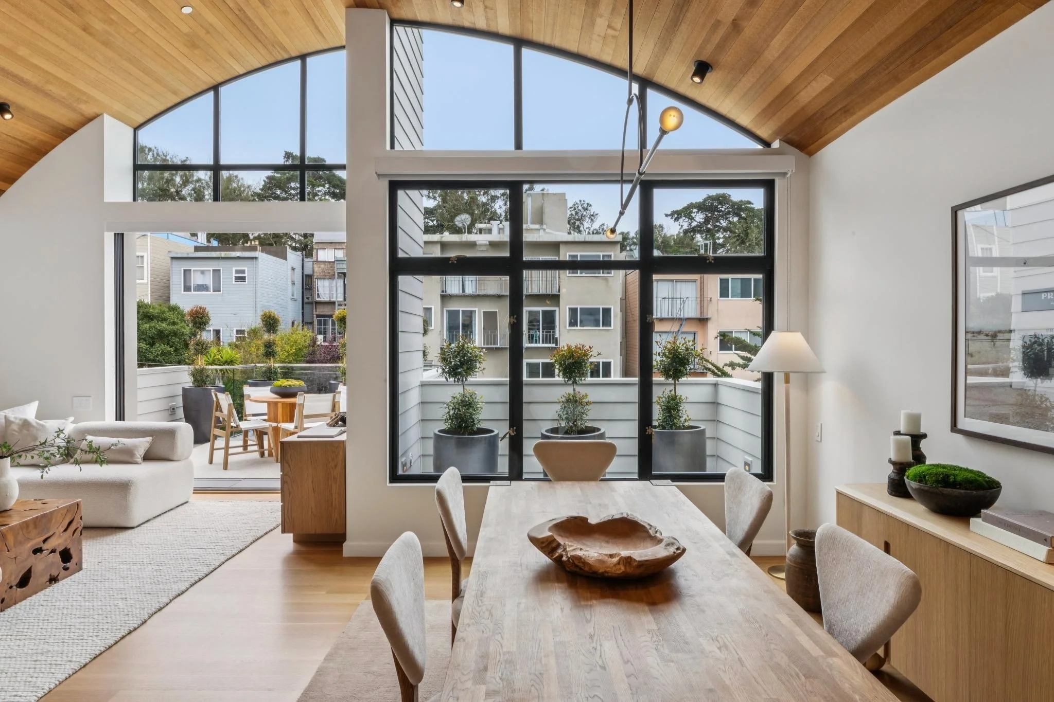 Open living room and dining area with large windows, wood ceiling, and modern furniture including a dining table with chairs, a sofa, and potted plants.