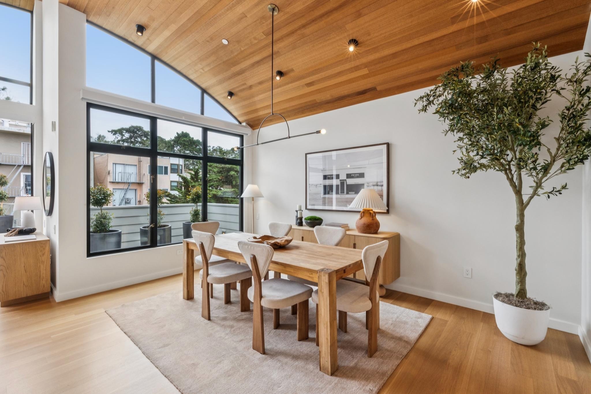 Modern dining room with a wooden table, six white chairs, large windows, potted plant, framed artwork, and pendant lighting.