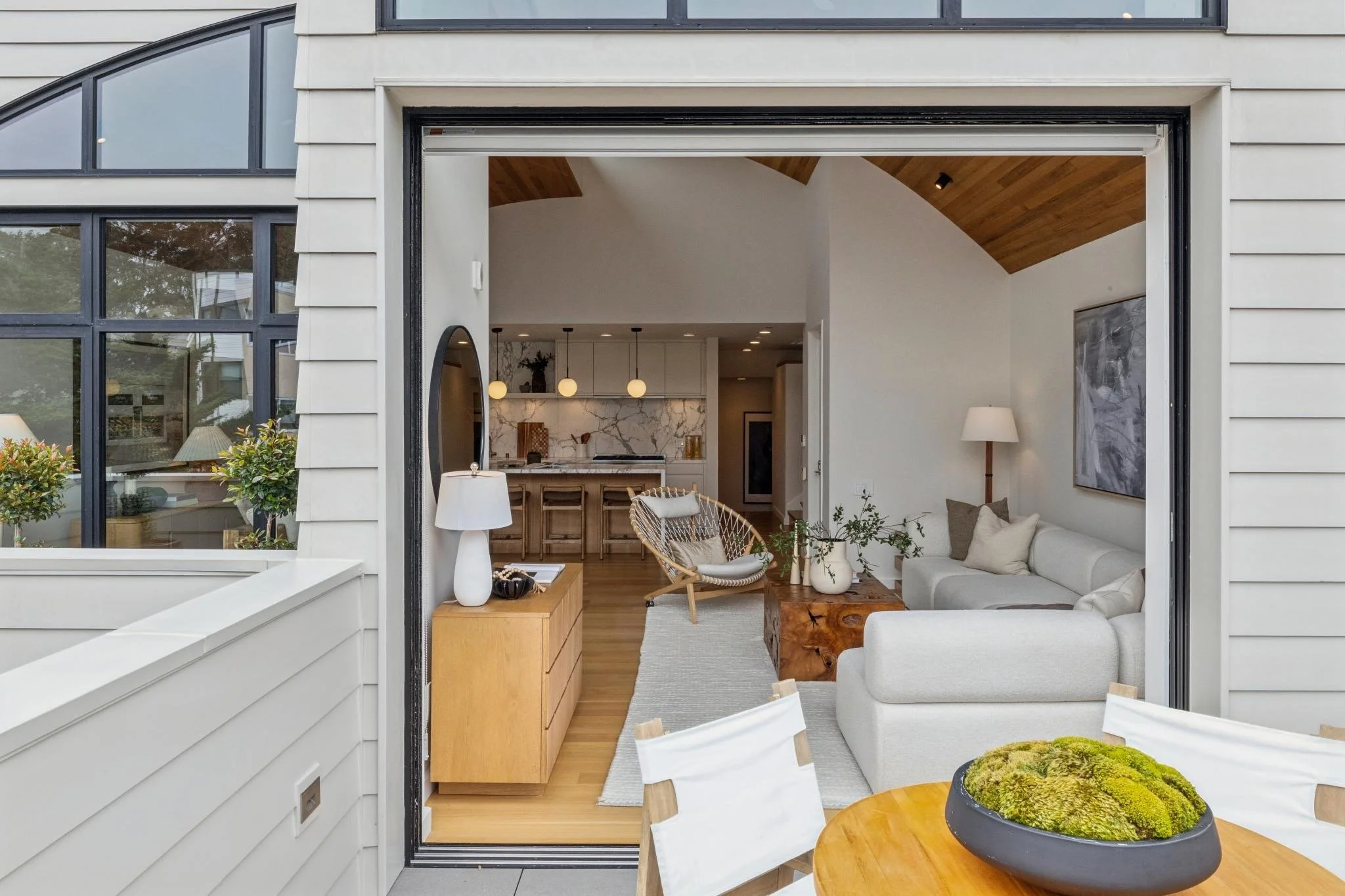 Modern home interior seen through large open sliding glass door, featuring white sofa, wooden coffee table, and a dining table with a plant in a black pot.