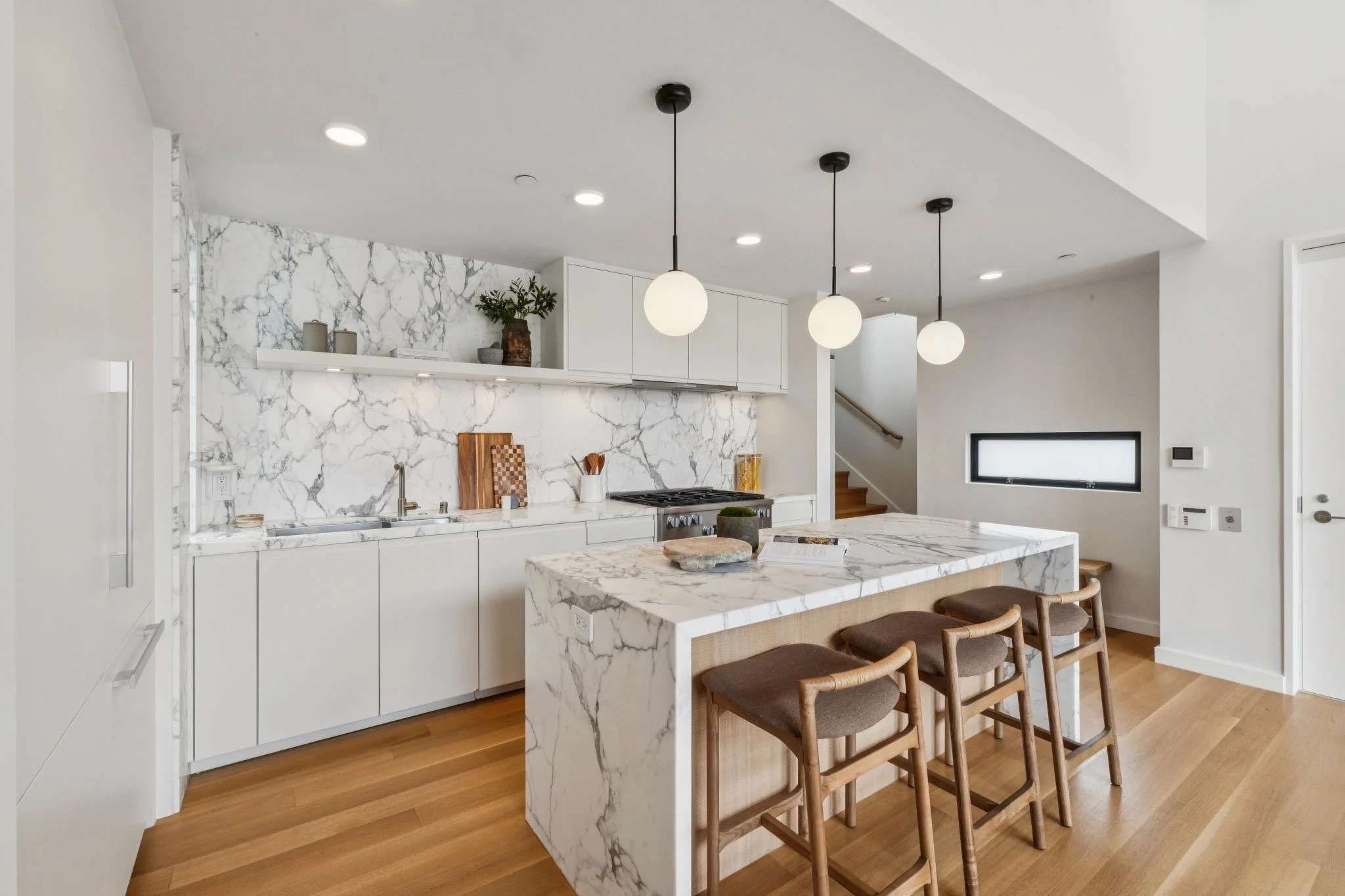 Modern kitchen with white marble countertops and backsplash, white cabinets, a kitchen island with a marble top, wooden bar stools, and black pendant lights, with wooden flooring and a small window.