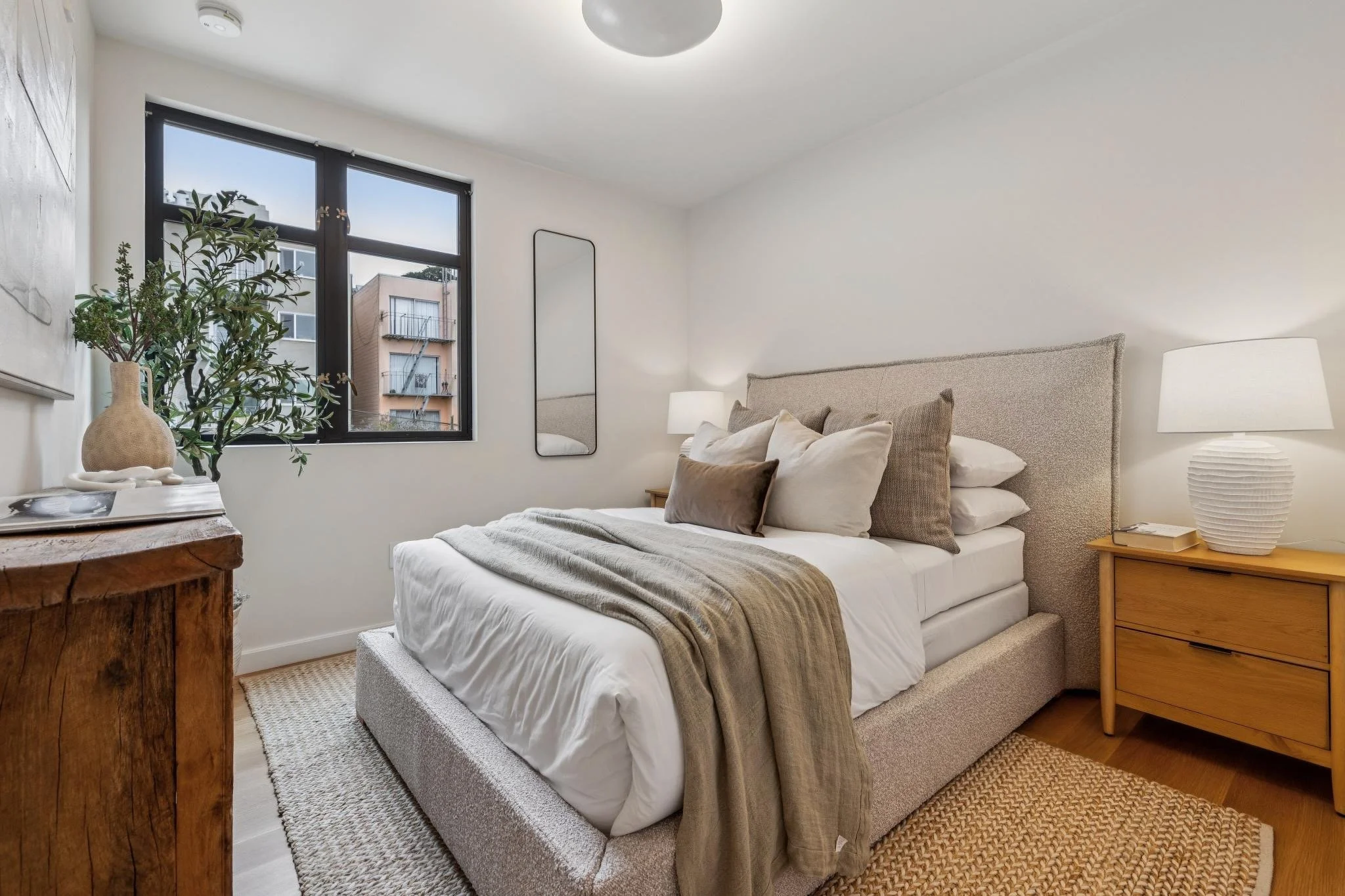 A bedroom with a bed, plant, mirror, and nightstand; natural light coming through a window.