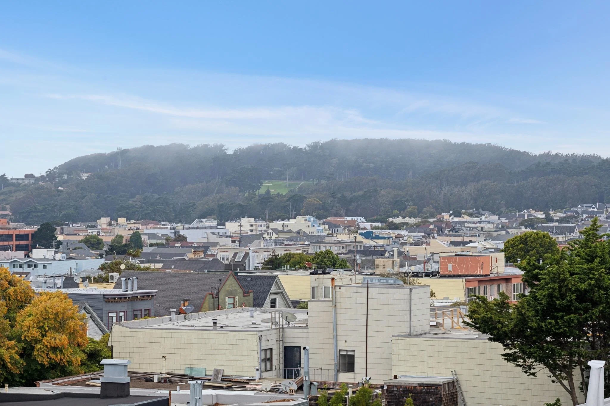 A cityscape with residential buildings in the foreground, a hilly green area in the background, under a clear blue sky.