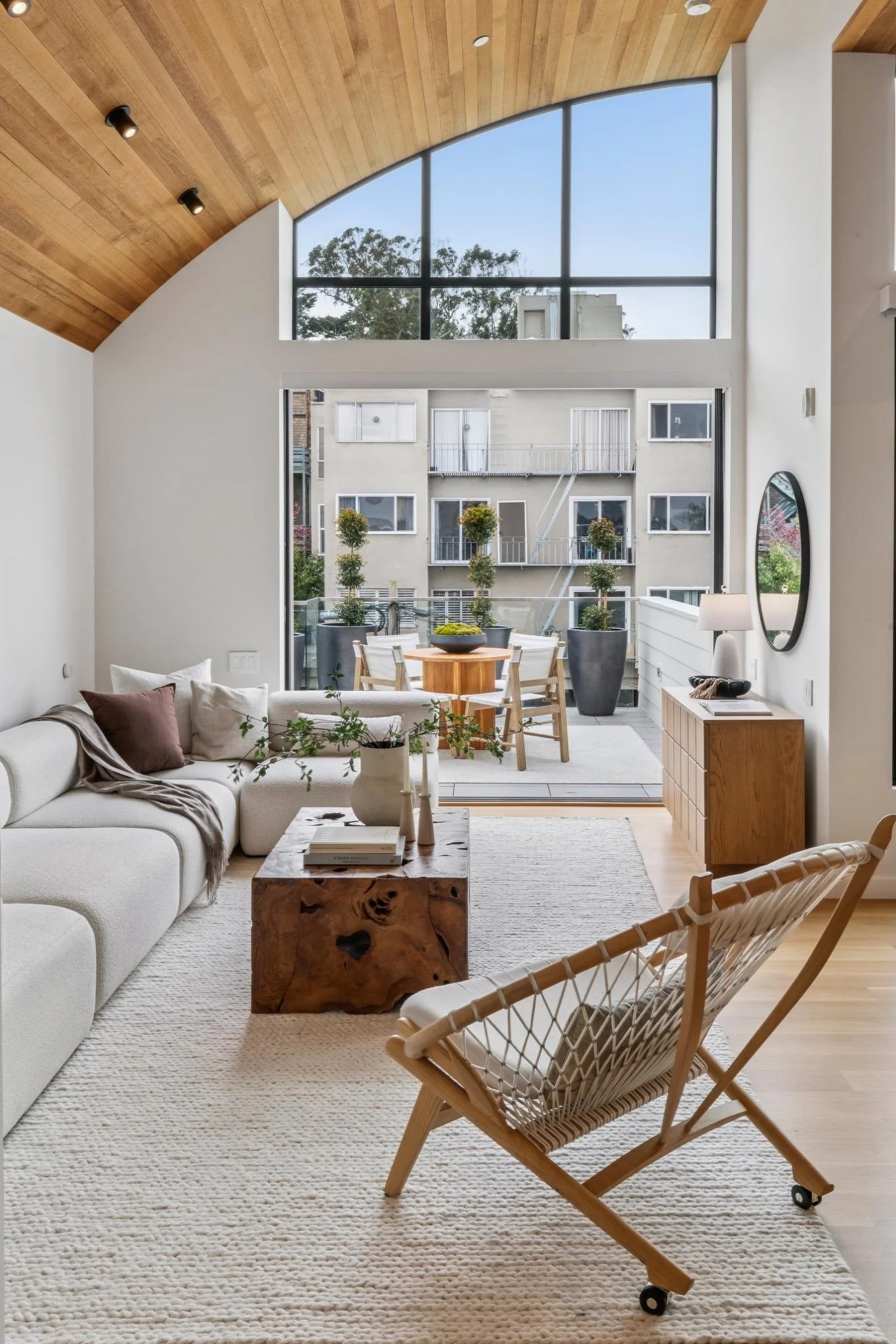 Living room with large window, white couch with pillows, wooden coffee table, and a woven armchair with wheels, cream-colored rug.