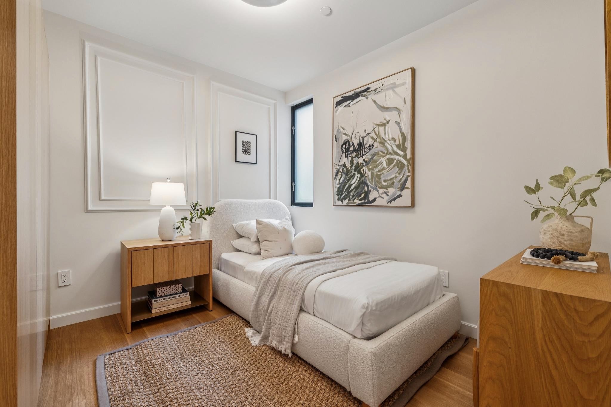 A bedroom with white walls, a small bed with a beige blanket and white pillows, a wooden nightstand with a white lamp and a plant, three abstract artworks on the walls, a window, a wicker rug on wooden flooring, and a wooden dresser