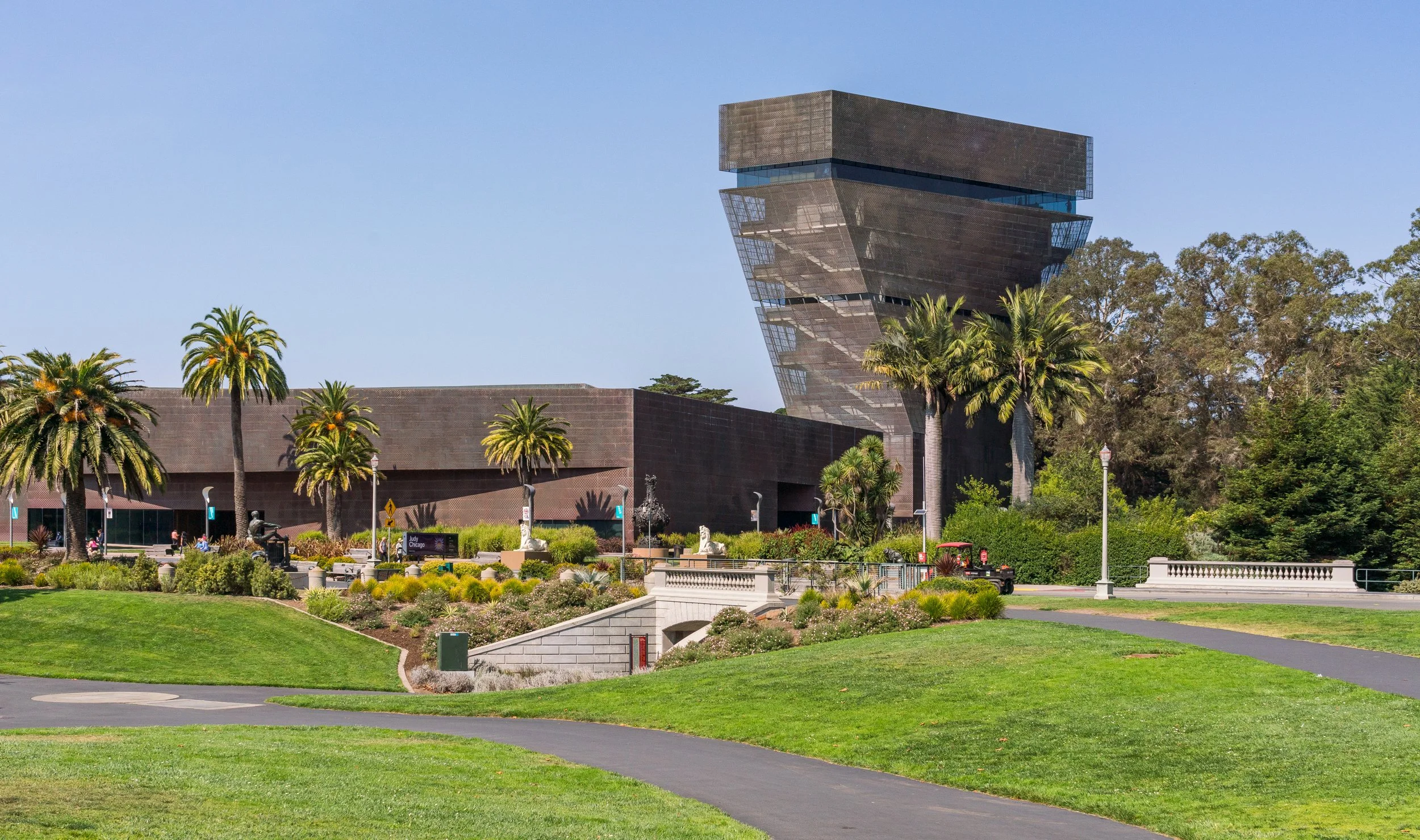 The modern black building with a tilted, stacked design is surrounded by palm trees, green lawns, flower beds, and walkways under a clear blue sky.