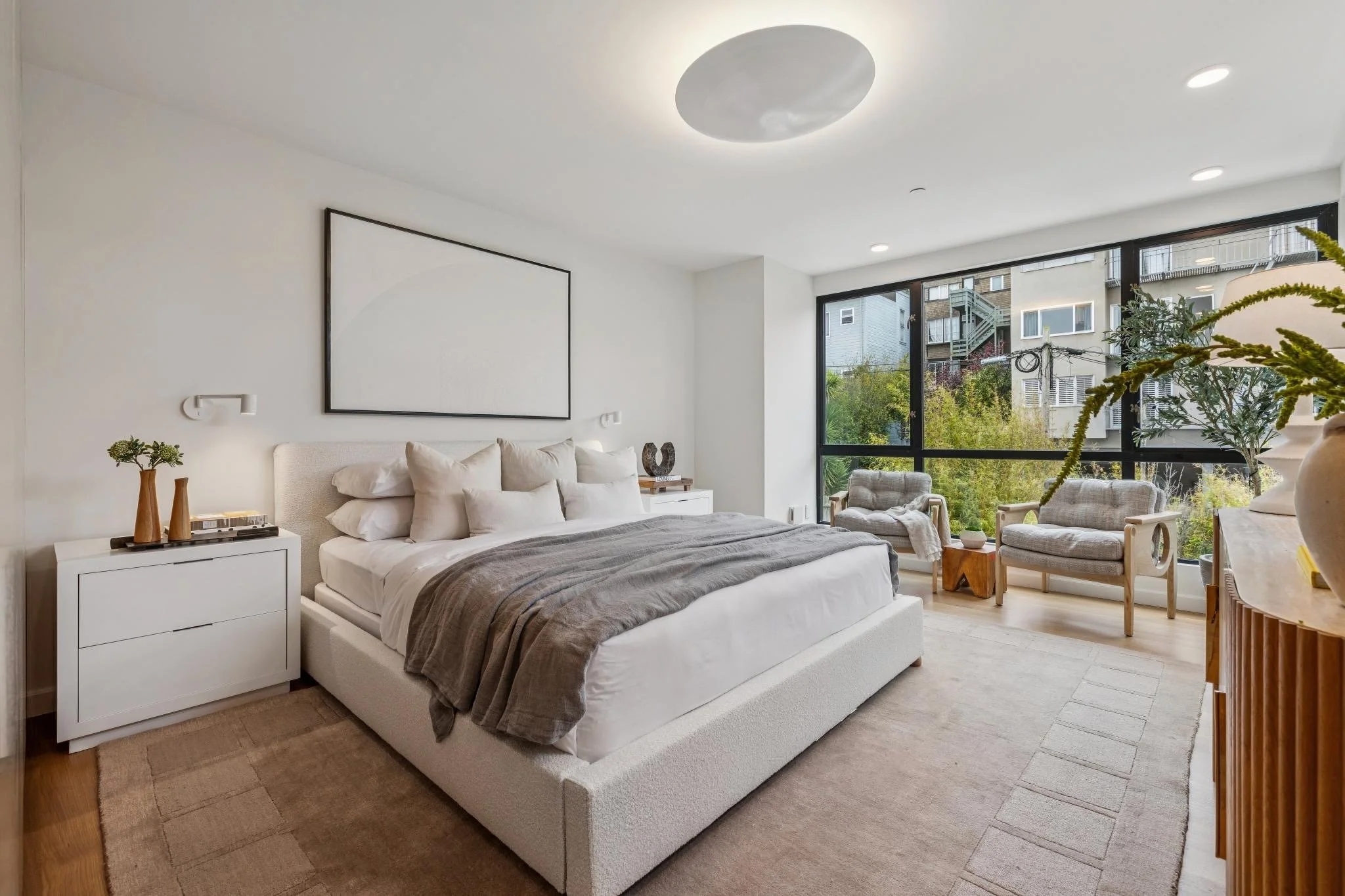 Modern bedroom with white bed, dark gray blanket, large window with city view, sitting area with chairs, and decorative plants.