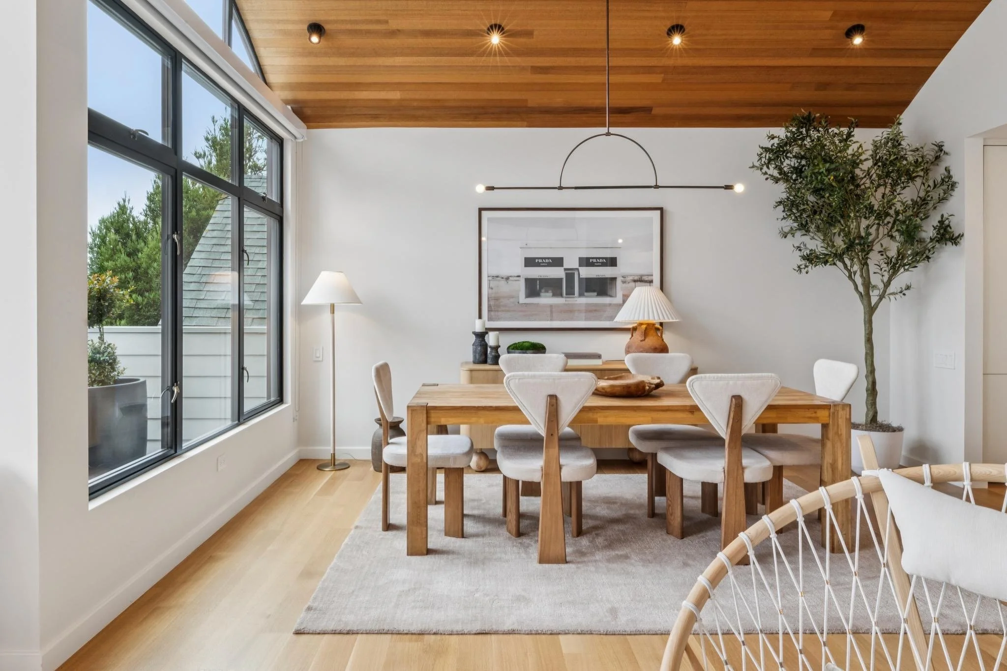 Modern dining room with large window, wooden ceiling, and minimalistic decor. A kitchen is visible in the background with a large framed photo on the wall, circular and linear light fixtures, a wooden dining table, and white chairs. Several decorativ