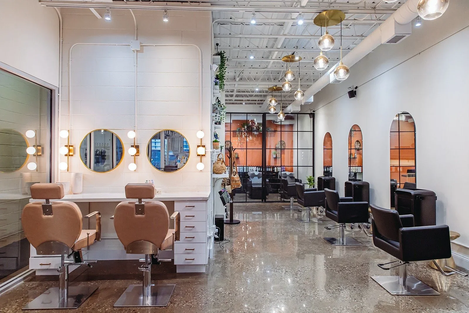 Modern hair salon with beige and black styling chairs, mirrors, and bright lighting, decorated with plants and hanging globes, facing a clean, polished floor.