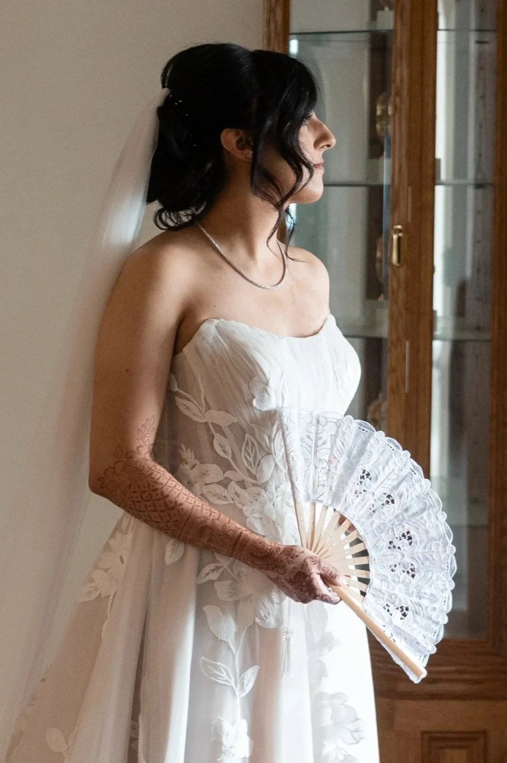 Woman in a wedding dress holding a lace fan, with henna designs on her arm, standing indoors near a wooden cabinet.