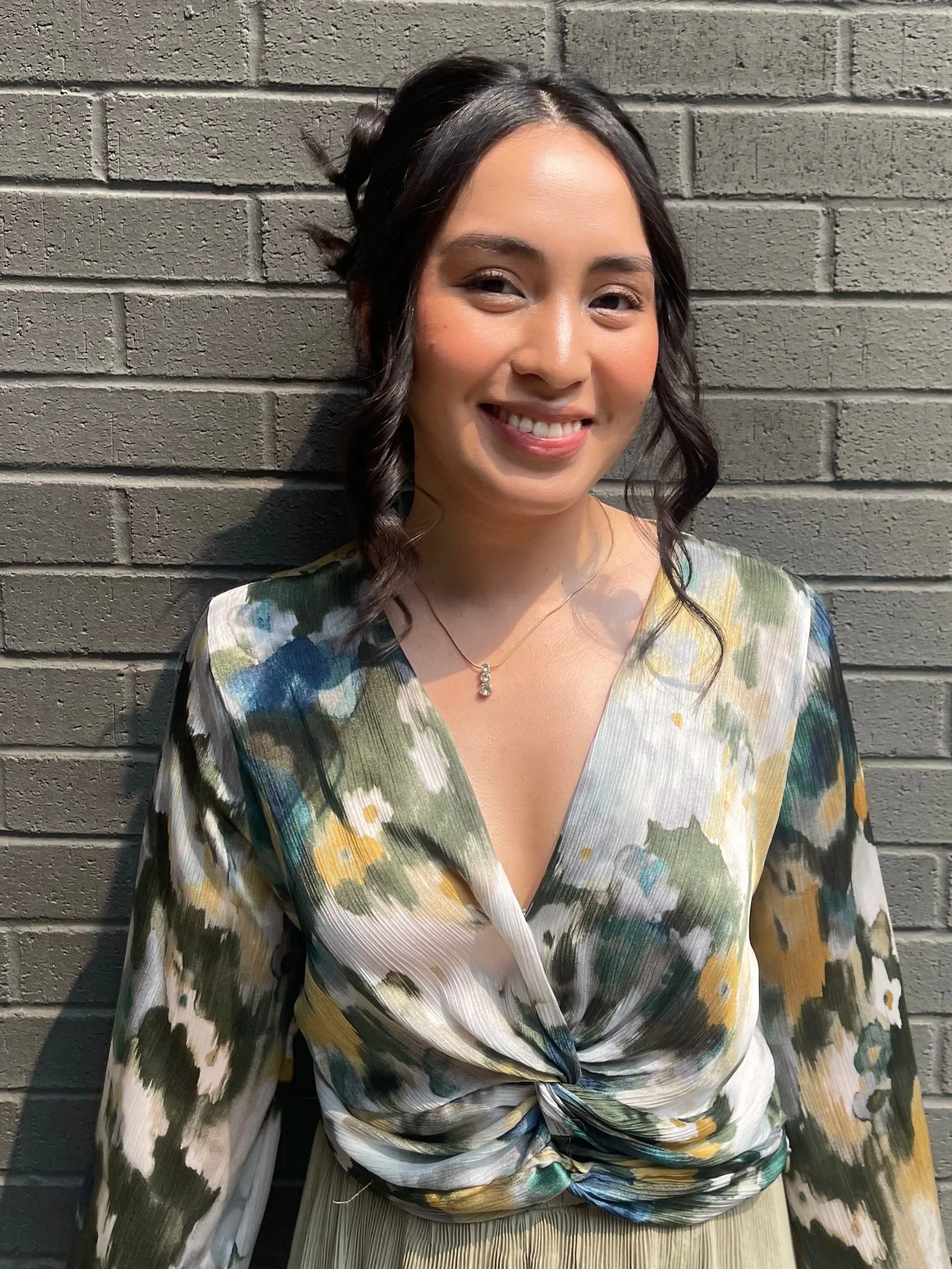 A woman with black hair styled in loose curls, wearing a pastel-colored, patterned blouse, smiling in front of a gray brick wall.