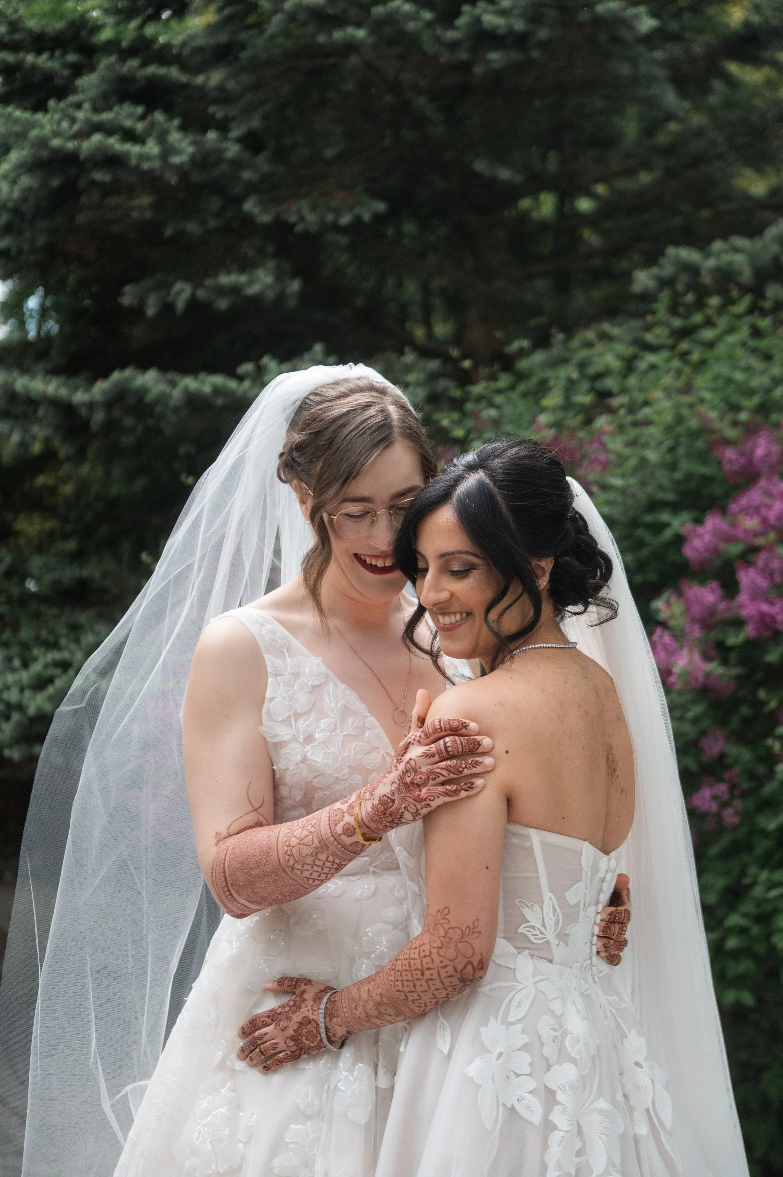Two women dressed in wedding gowns sharing a joyful moment outdoors, with one woman wearing a veil and the other adorned in a backless gown, surrounded by lush green foliage and purple flowers in the background.