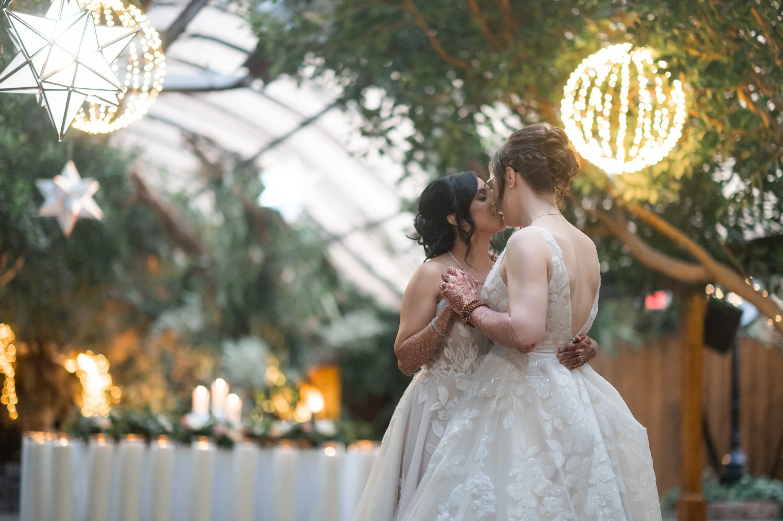 Two women in wedding dresses sharing a kiss at an outdoor wedding reception with string lights and hanging star-shaped lanterns.