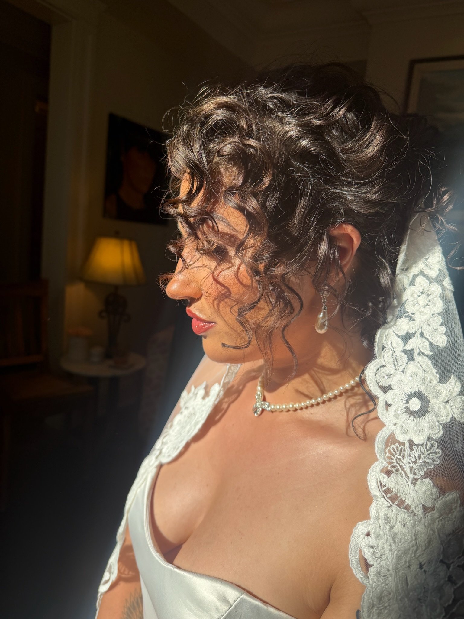 Close-up side view of a woman with curly brown hair, wearing a wedding dress with lace details on the shoulder and a pearl necklace with a bow pendant.