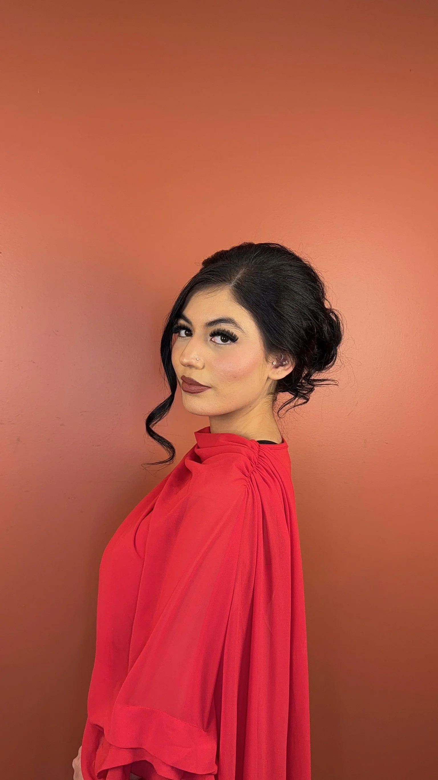 A woman with black hair styled in an updo with loose curls, wearing makeup, a nose piercing, and a red dress, standing against a gradient pink background.
