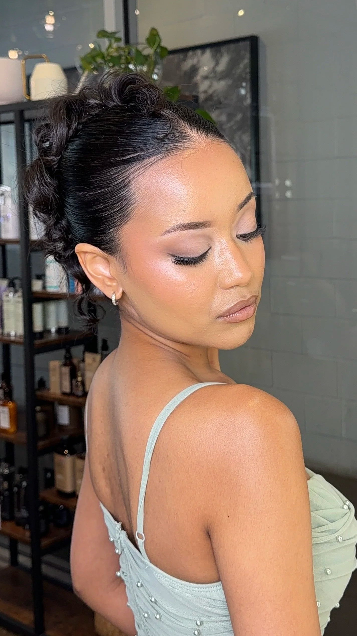 A woman with styled, wavy black hair, wearing makeup, earrings, and a light-colored spaghetti strap top with pearl embellishments, looking down with her eyes closed in a room with shelves and a gray wall.