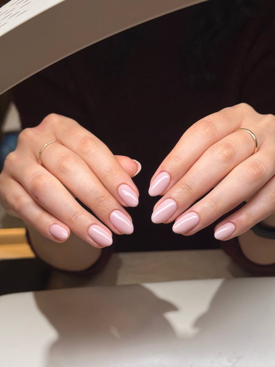 Close-up of well-manicured hands with light pink, glossy nail polish and thin gold rings on the fingers.