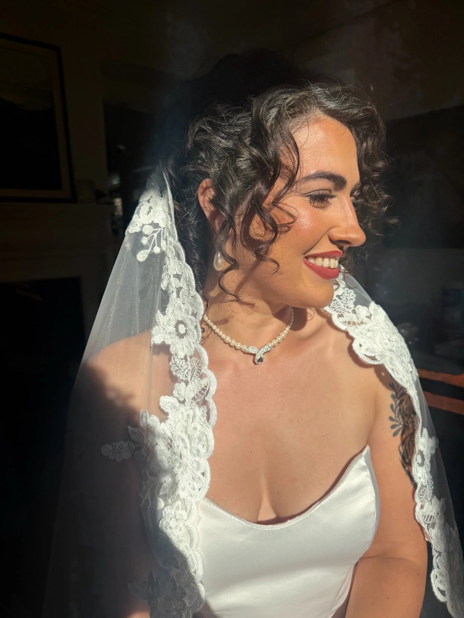 A woman with short, curly black hair and medium skin tone wearing a white satin dress with lace trim and a pearl necklace. She is smiling and appears to be dressed for a wedding or special occasion.