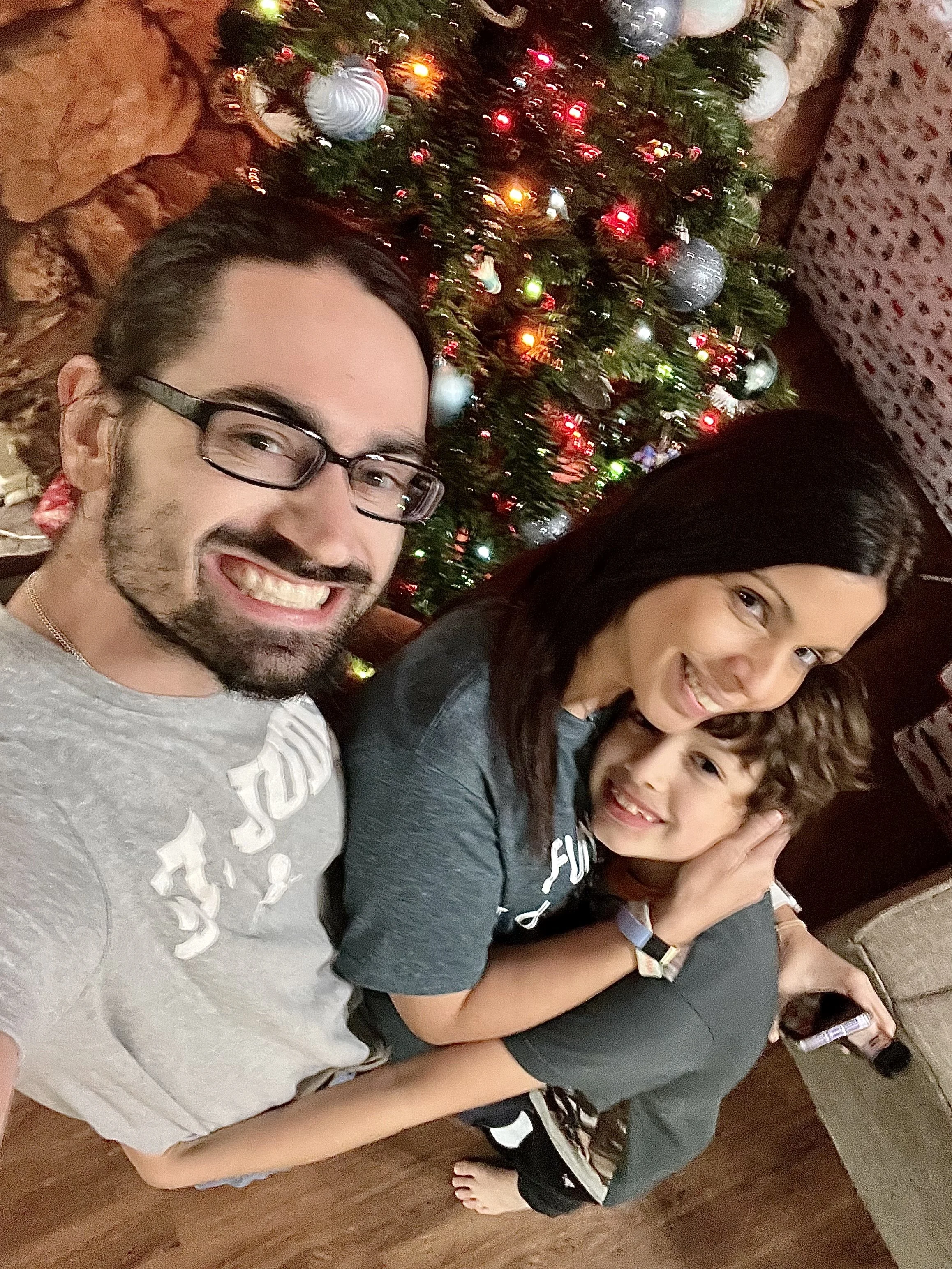 A group of people gathered around a Christmas tree decorated with white baubles and colorful lights. Two women and a young boy are smiling and posing for the photo. The person on the left is a man with glasses, a beard, and a gray shirt. The person next to him is a woman with dark hair and glasses. The boy has curly hair and is smiling with his hand on the woman's shoulder. In the background, there is a brick wall and other people partially visible.