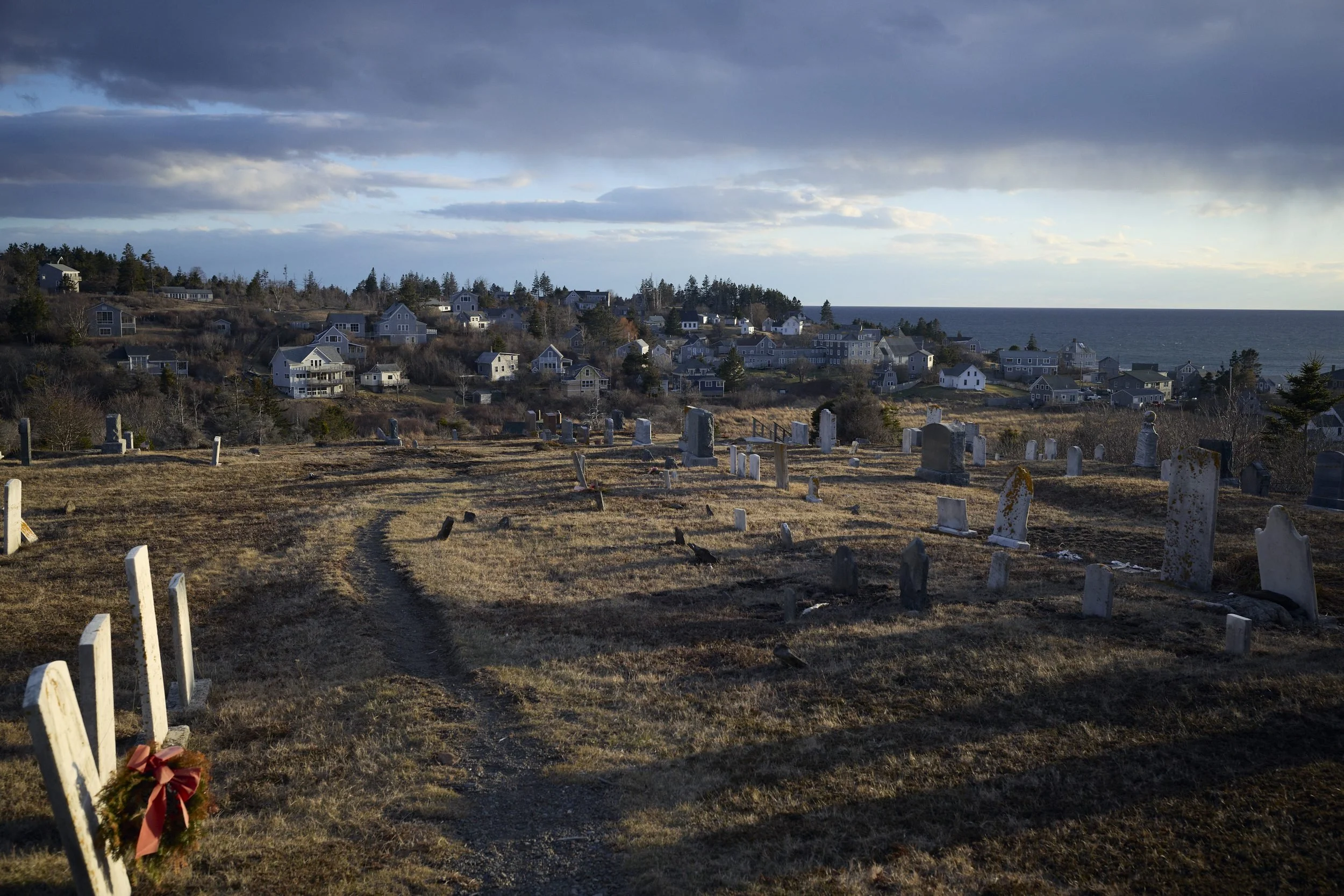 A cemetery on a hillside overlooks a coastal town with houses and evergreen trees under a cloudy sky.