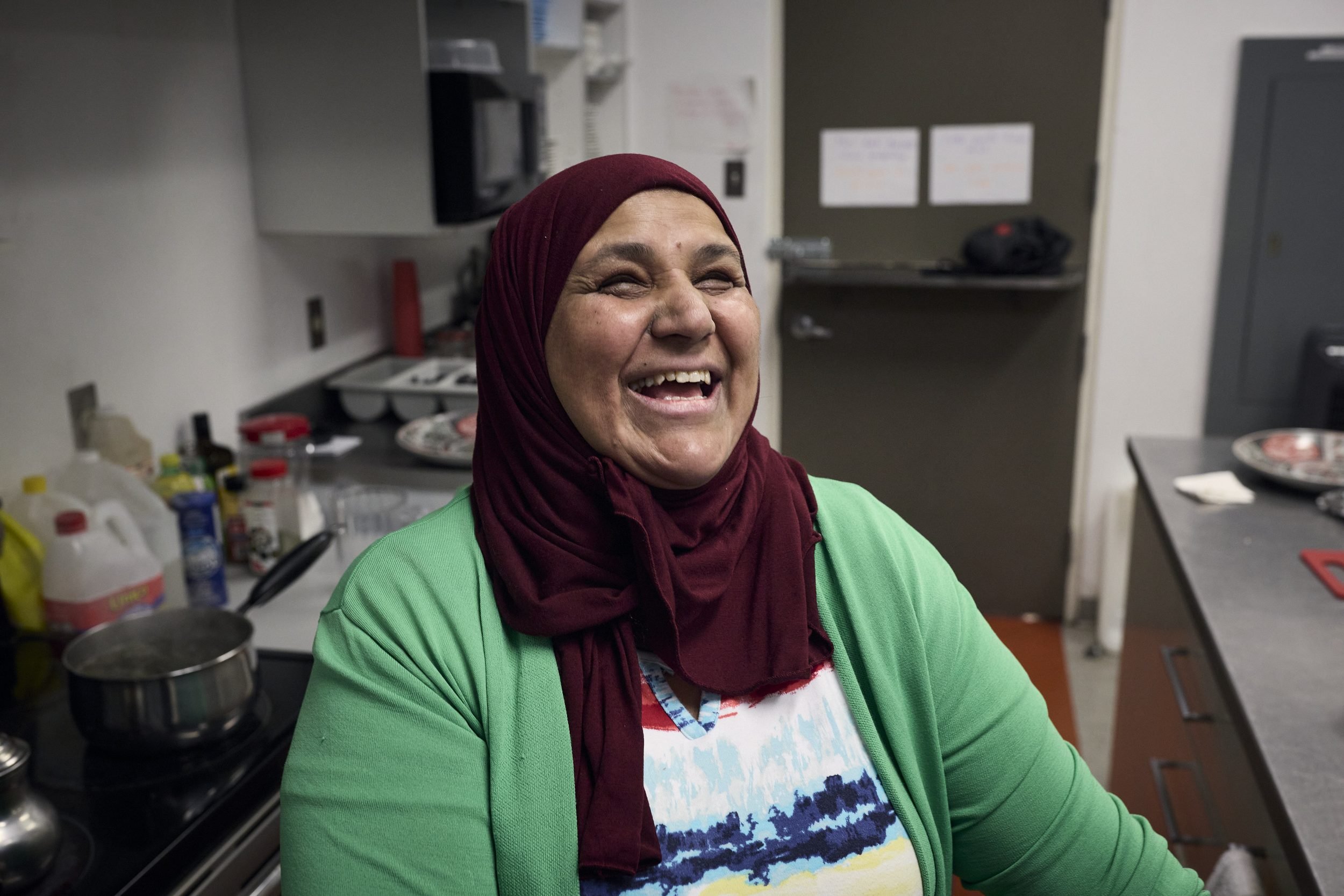 A woman wearing a burgundy hijab and green cardigan laughs in a kitchen.