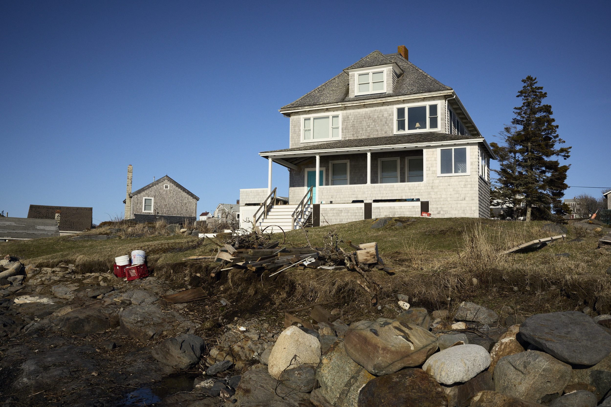 A weathered three-story house sits on a rocky coastal shore with driftwood and debris scattered across the beach.