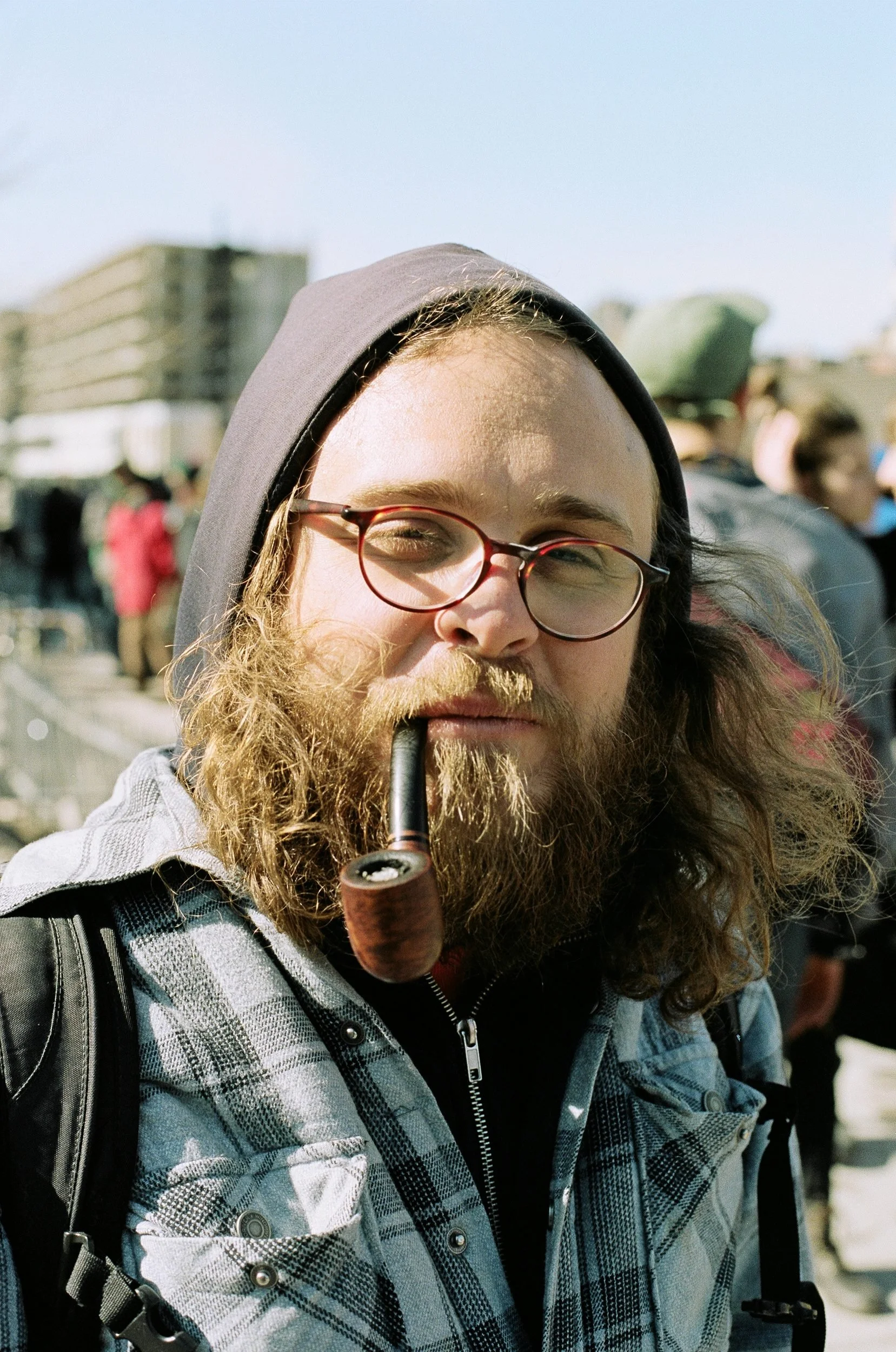 Man with long beard and glasses smoking a pipe, wearing a hoodie and plaid shirt outdoors.