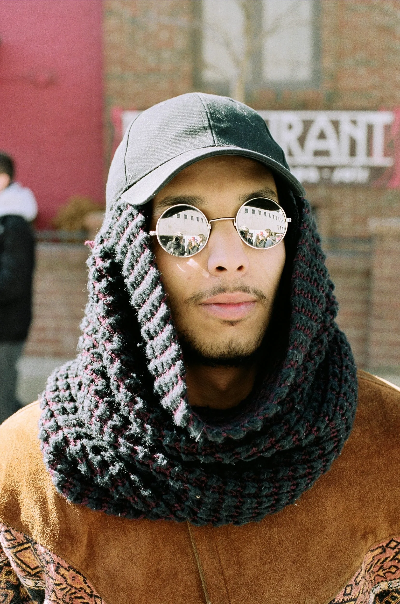 Person wearing round glasses, gray cap, and knit scarf over brown jacket stands against brick building backdrop.